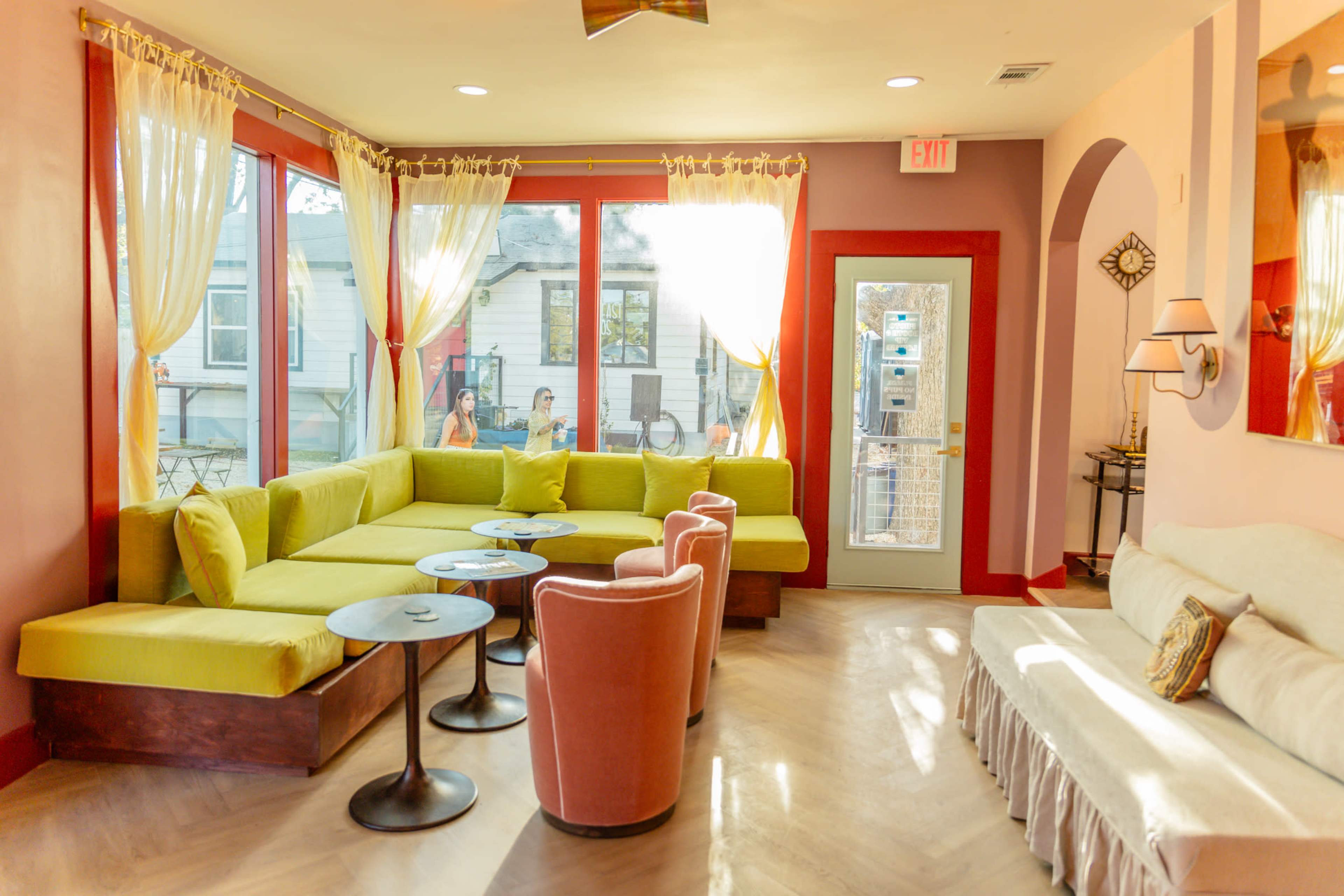A bright, modern seating area features a large green sectional, pink chairs, and round tables, surrounded by windows with sheer curtains.