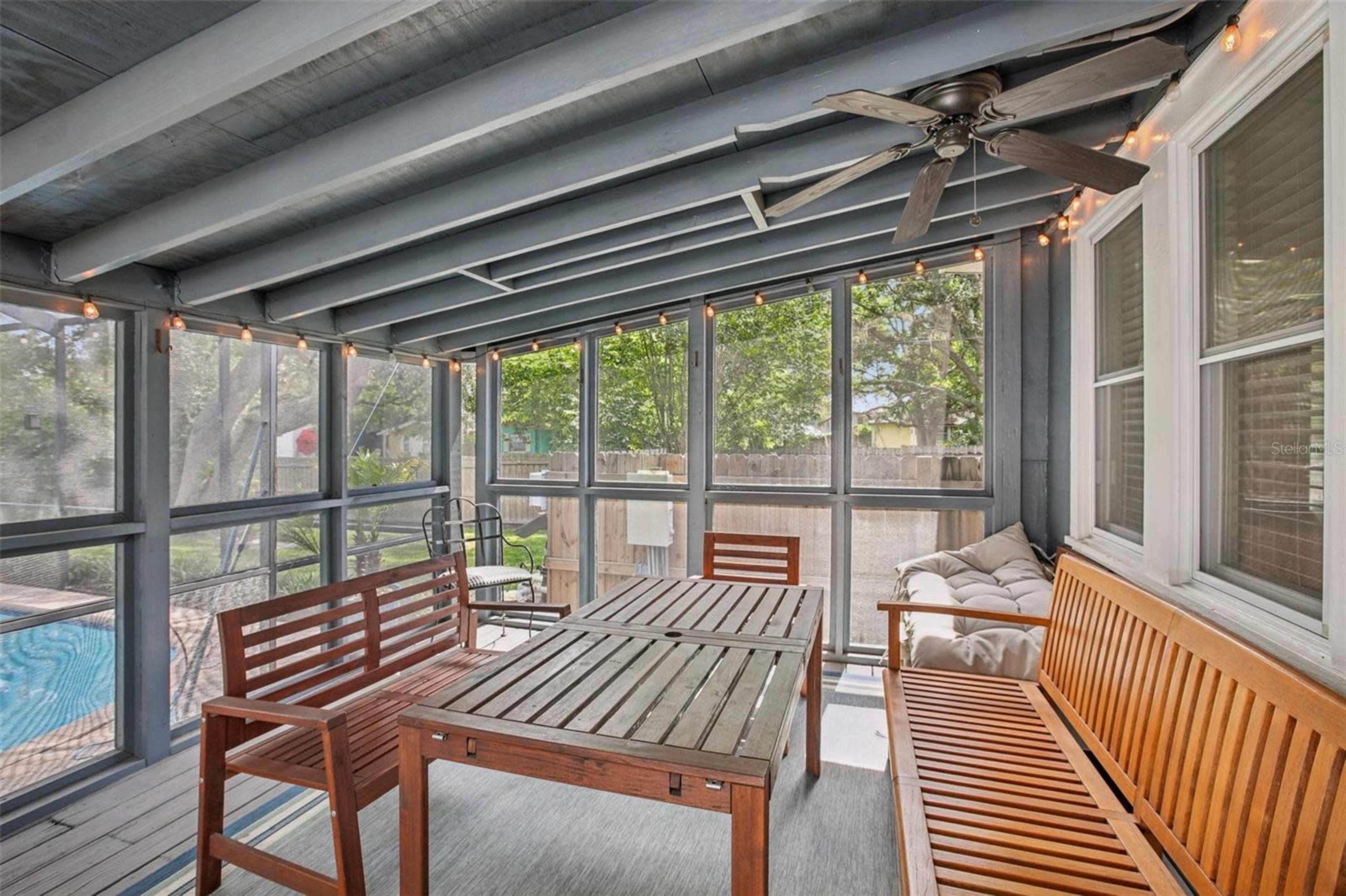 The image shows a screened-in porch featuring wooden furniture, including a table and benches, with ceiling fans and string lights above.