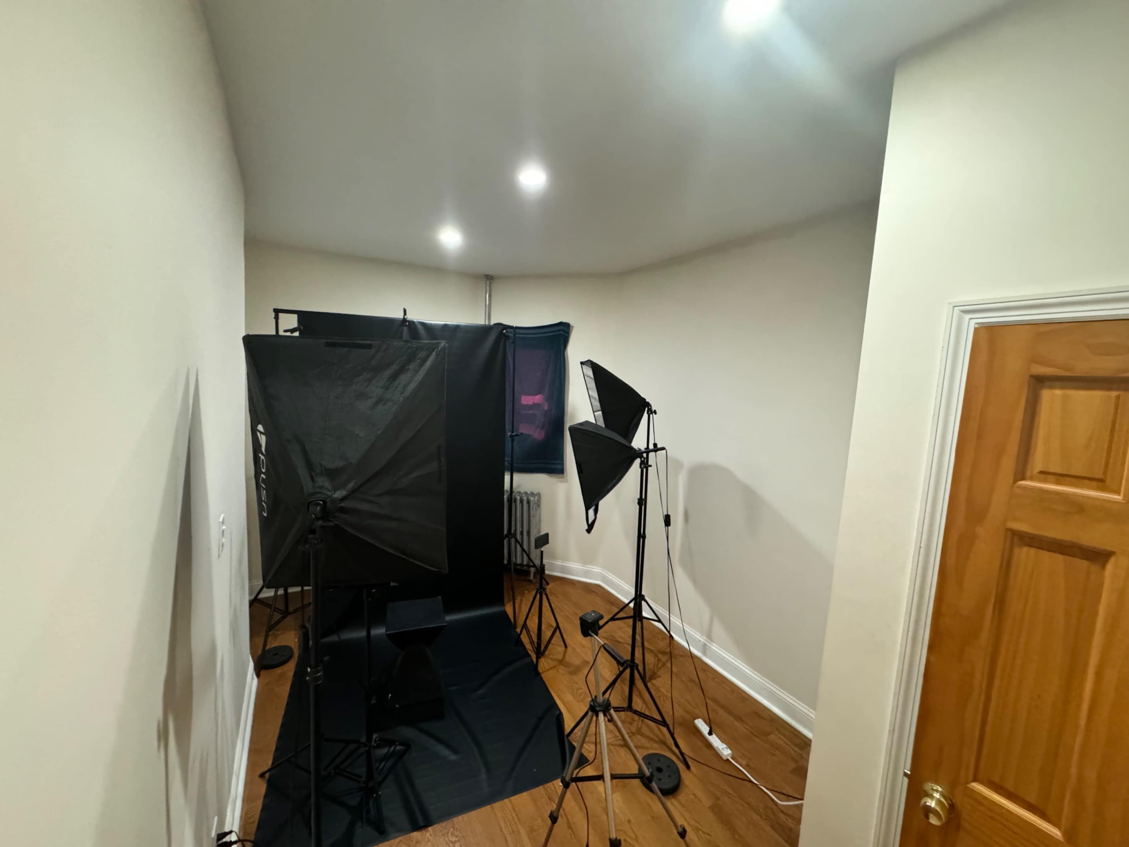 The image shows a photography setup in a small room, featuring softboxes, a backdrop, and various equipment arranged on a dark floor.