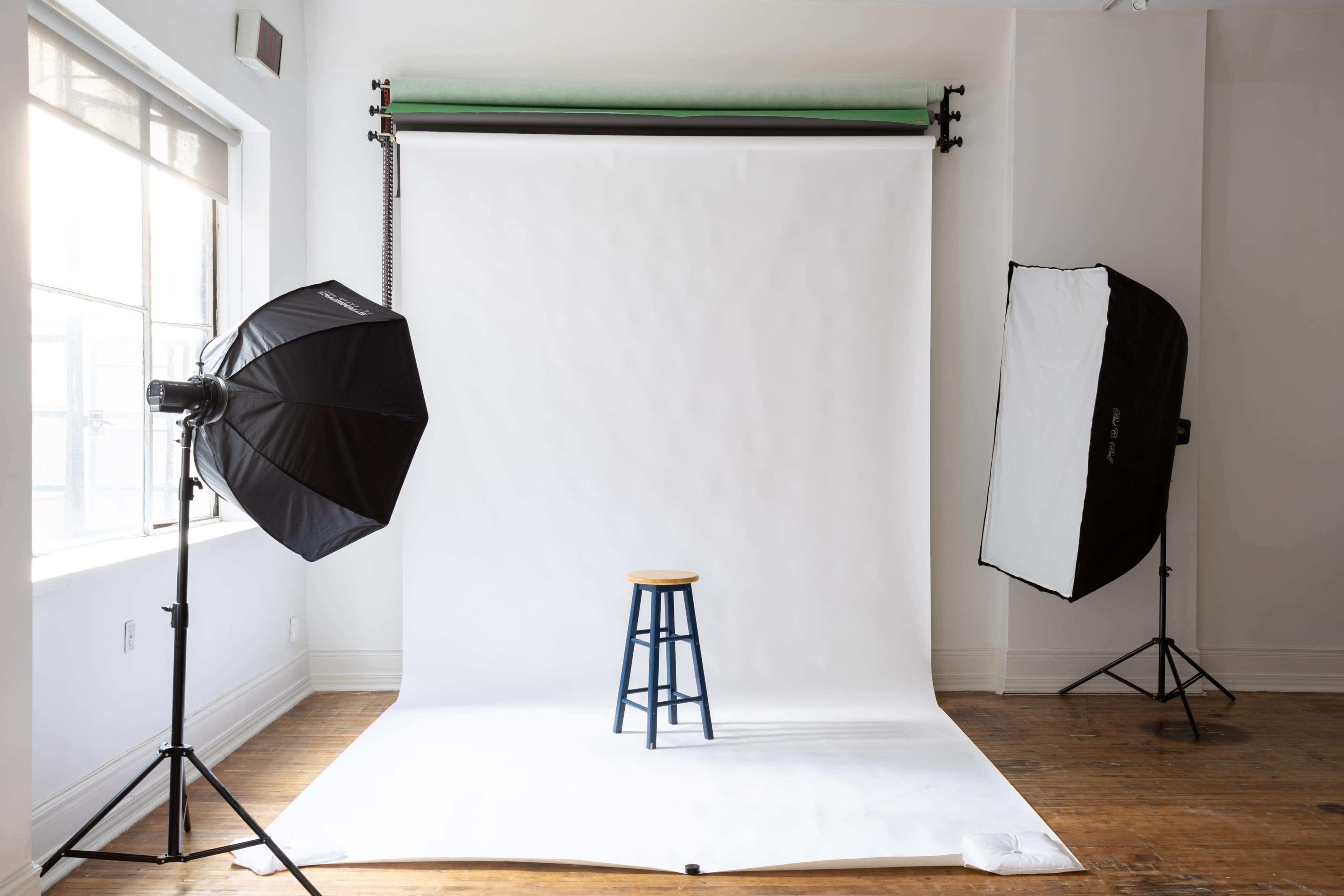 A photography studio with a white backdrop, two light sources, and a wooden stool in the center.