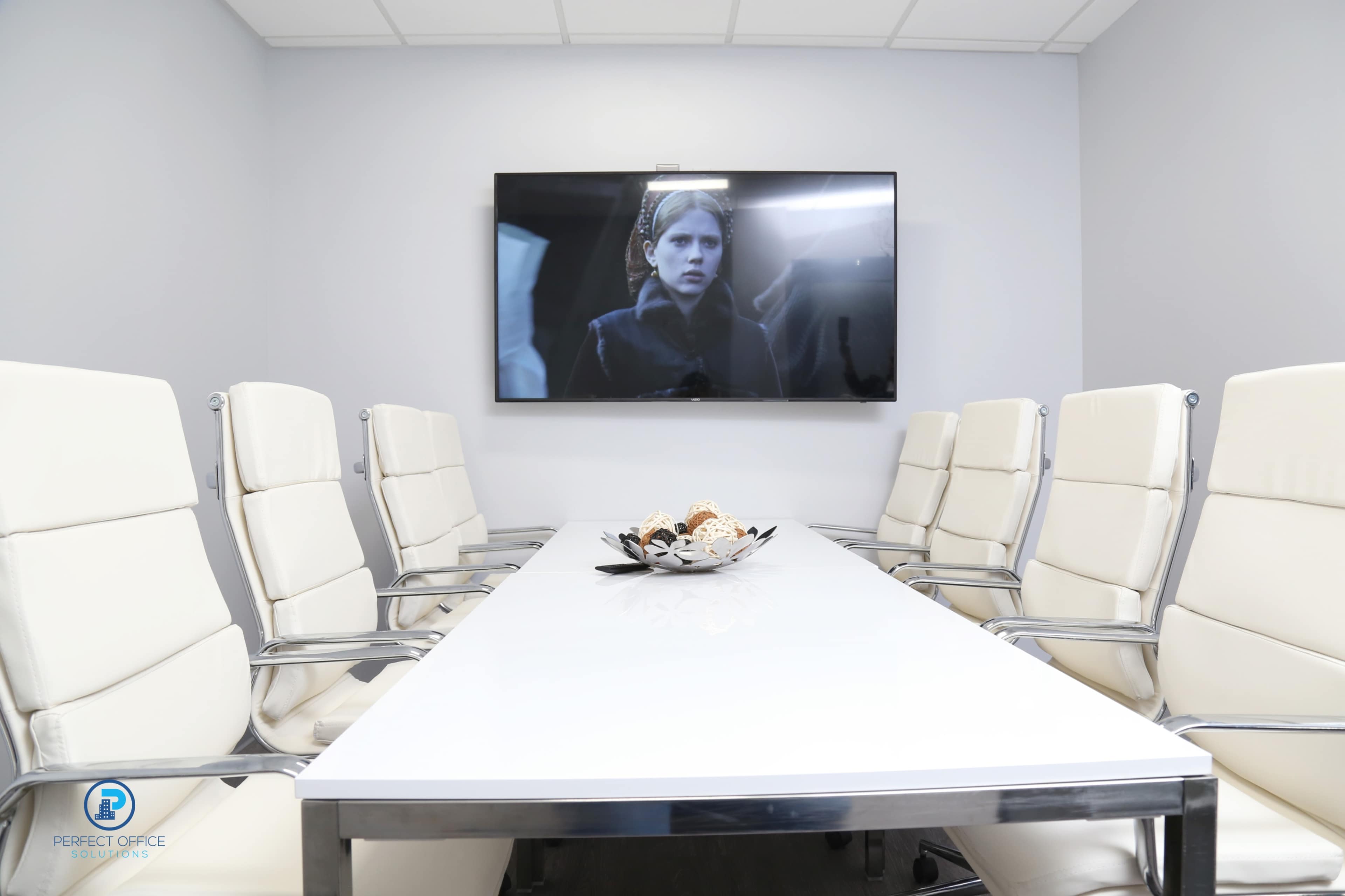 A modern conference room features a long white table surrounded by eight white chairs and a large television screen mounted on the wall.
