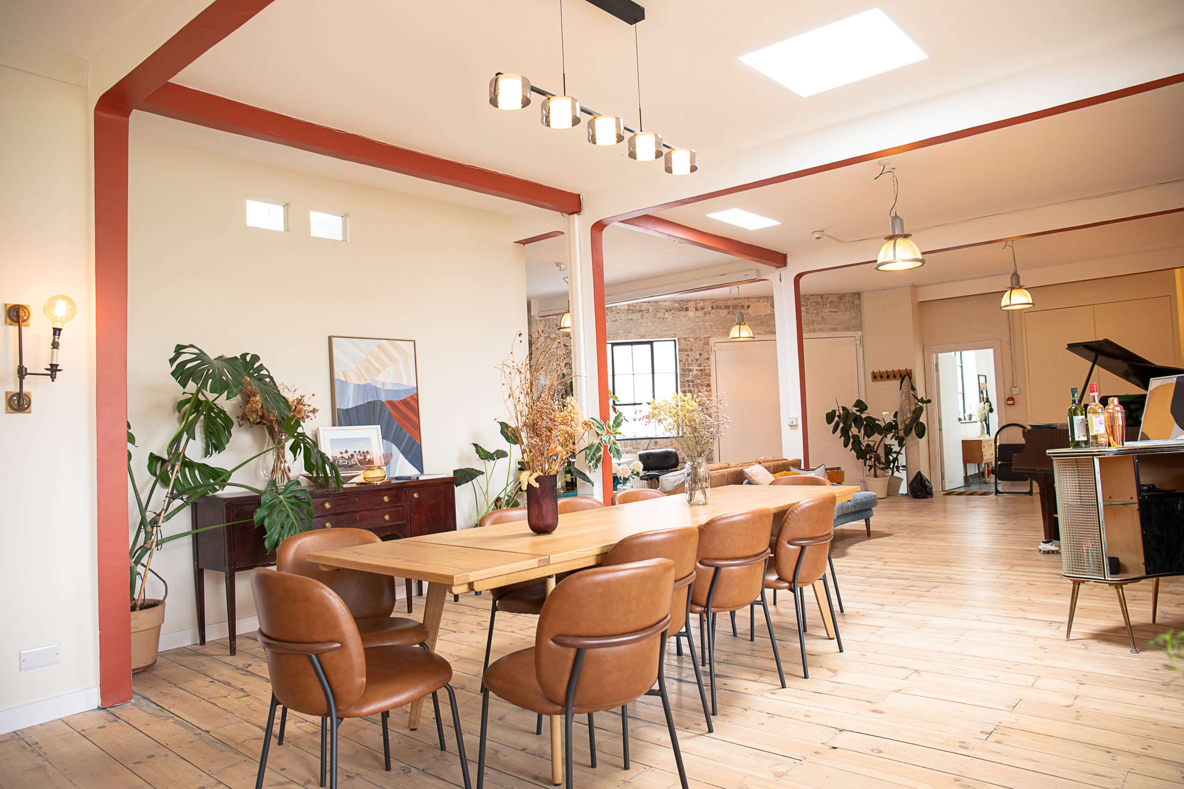 The image shows a spacious dining area with a wooden table surrounded by brown chairs, adjacent to a living space adorned with plants and a piano.