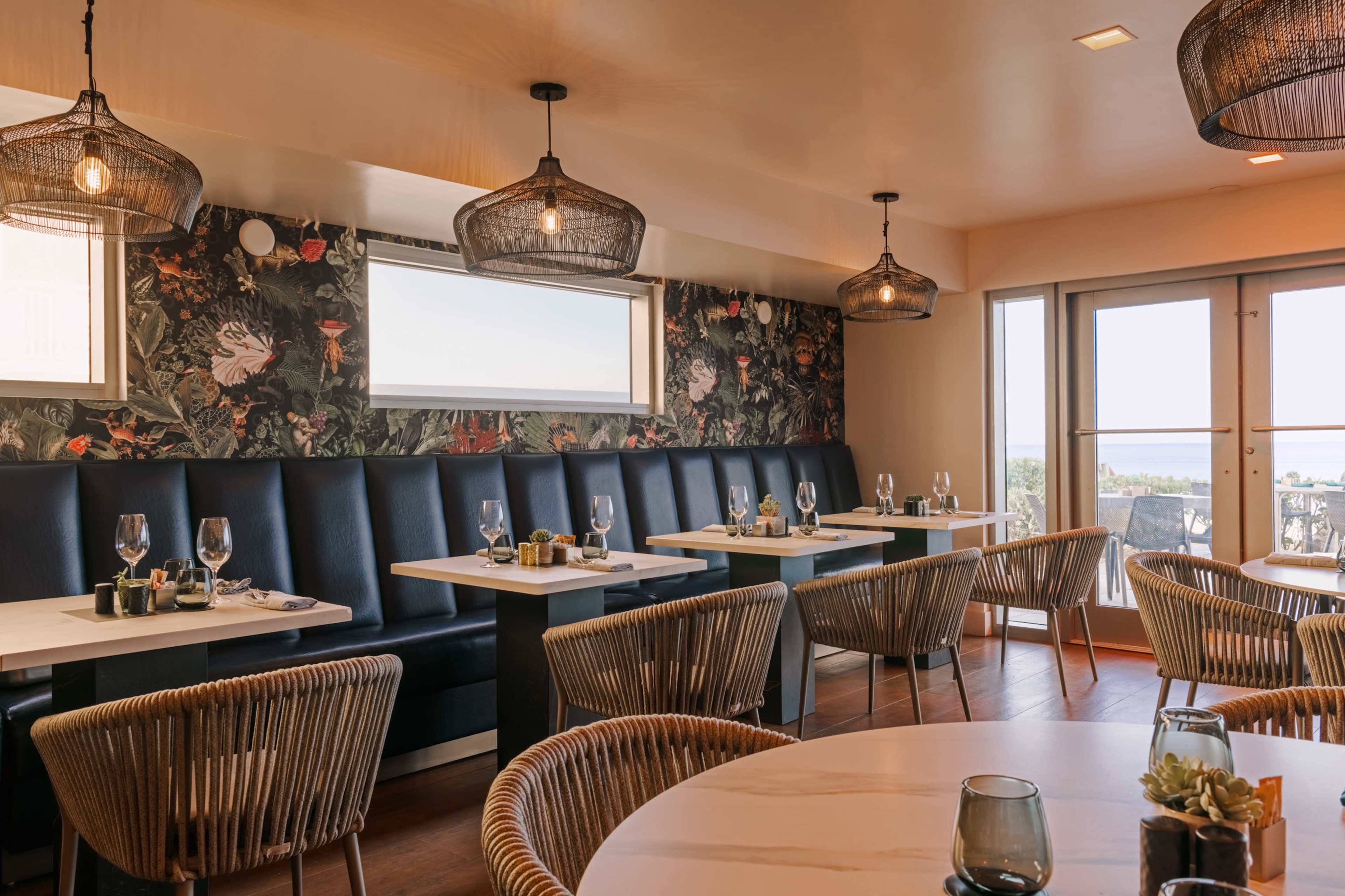 The image shows a modern restaurant interior featuring dark blue upholstered seating, wooden tables, and woven light fixtures, with large windows providing natural light.