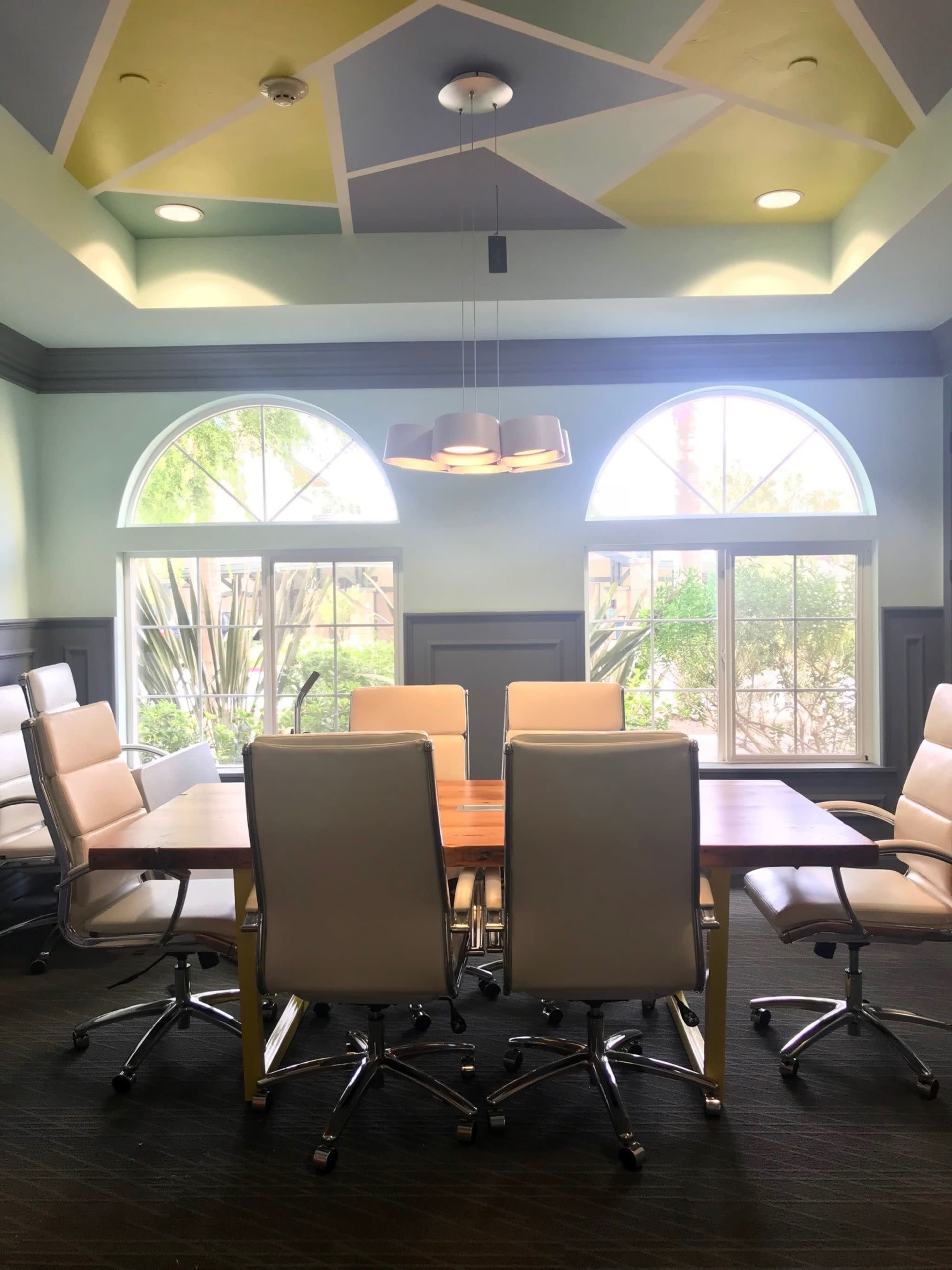 A conference room features a wooden table surrounded by rolling office chairs beneath a patterned ceiling and large windows.