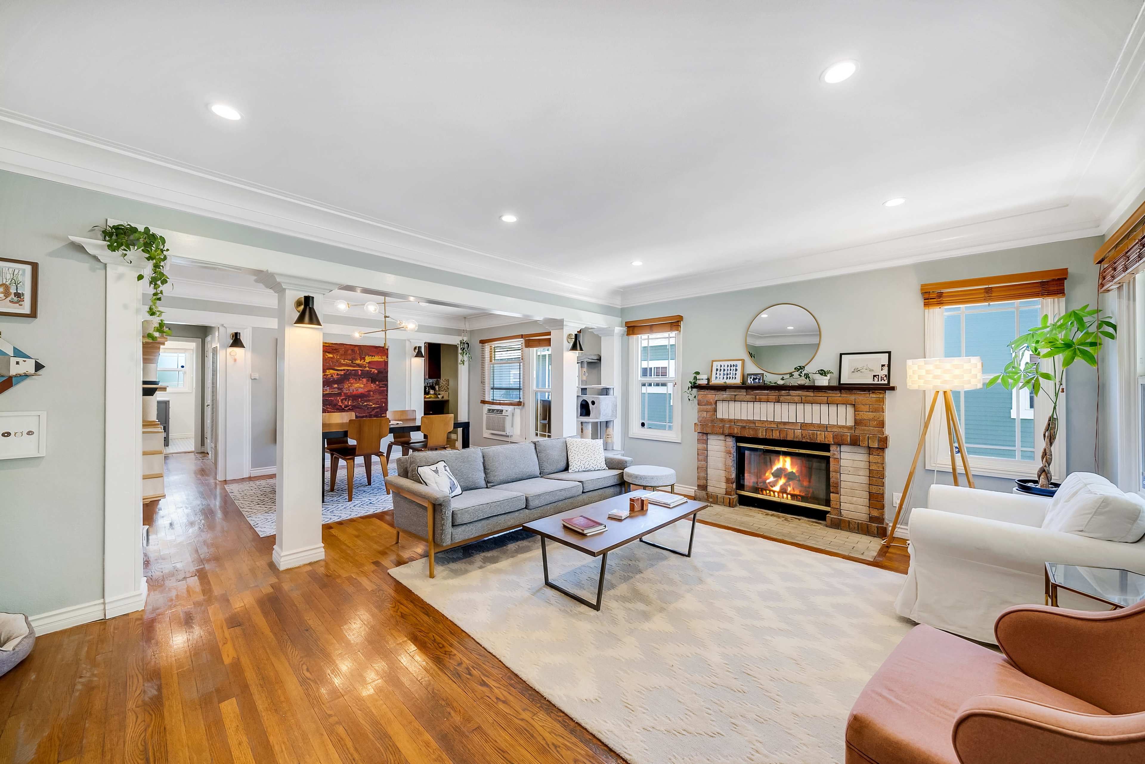 A cozy living room features a gray sofa and armchair, a brick fireplace, and hardwood floors, with a dining area visible through an open space.