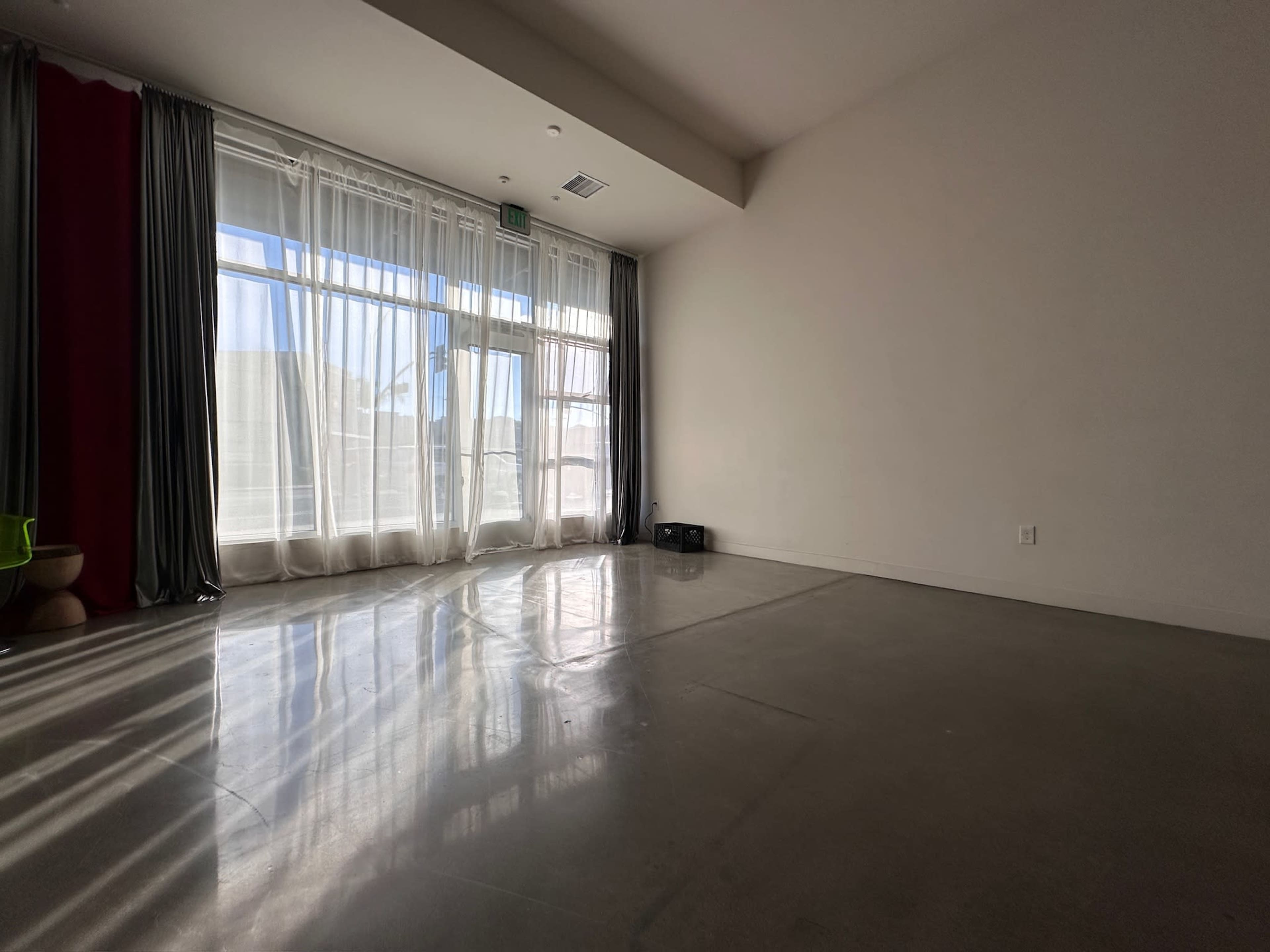 The image shows a spacious, minimally furnished room with large windows covered by sheer curtains, casting shadows on the polished concrete floor.