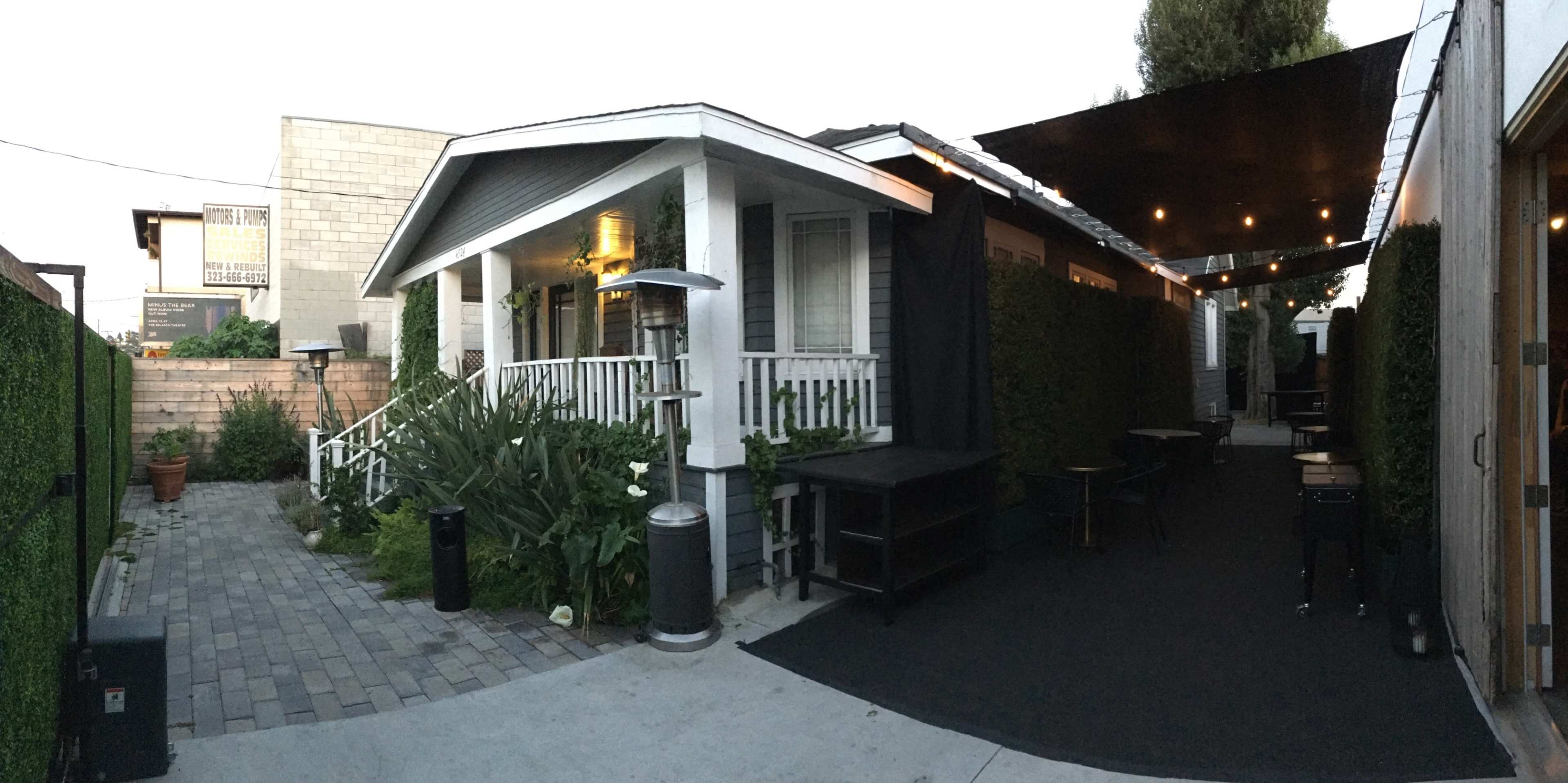 The image shows a narrow outdoor space featuring a house with a porch, surrounded by potted plants, black fabric awnings, and tables set up on a paved pathway.