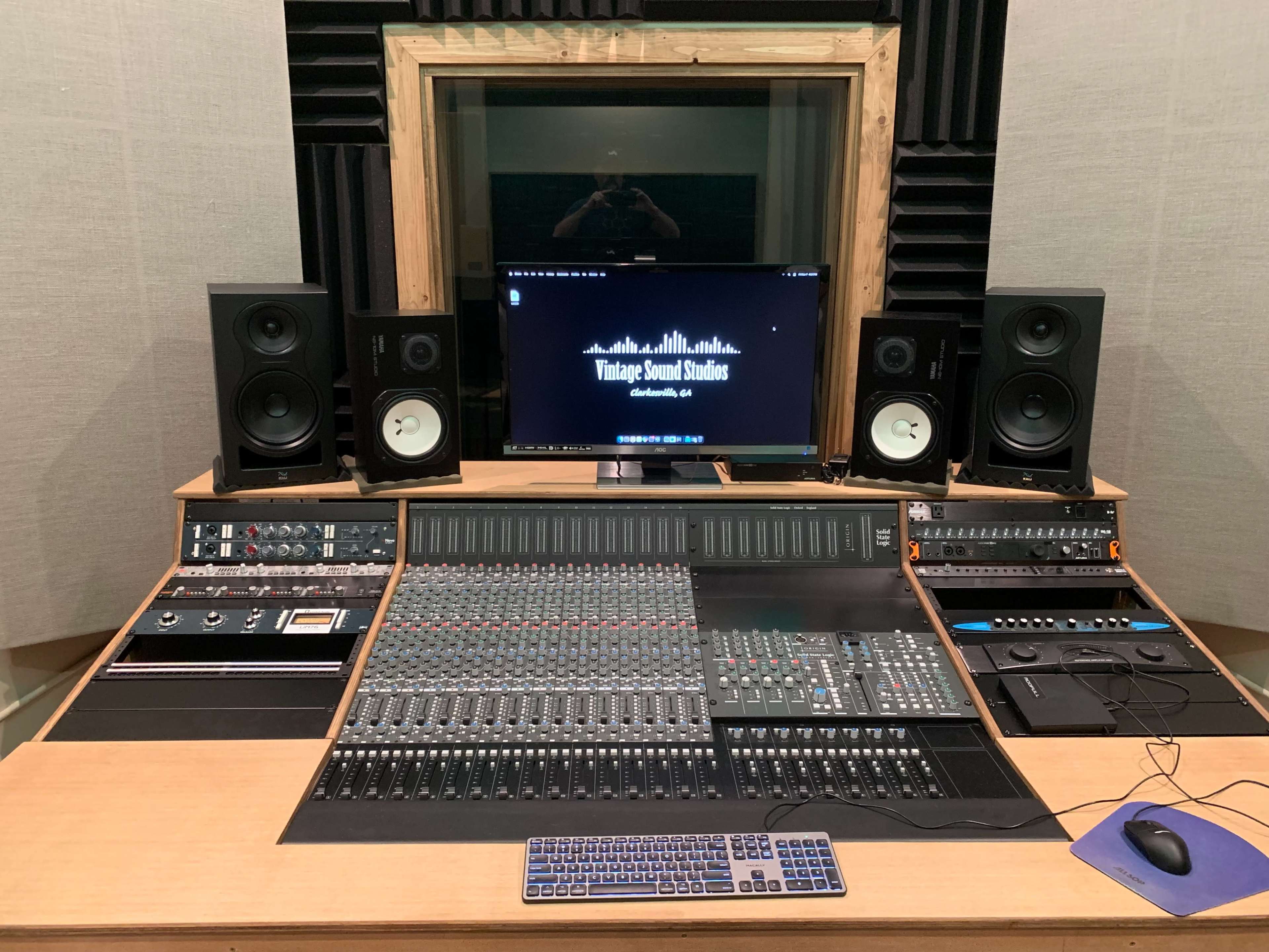 The image shows a professional audio mixing studio featuring a large mixing console, computer monitor, speakers, and soundproofing panels on the walls.