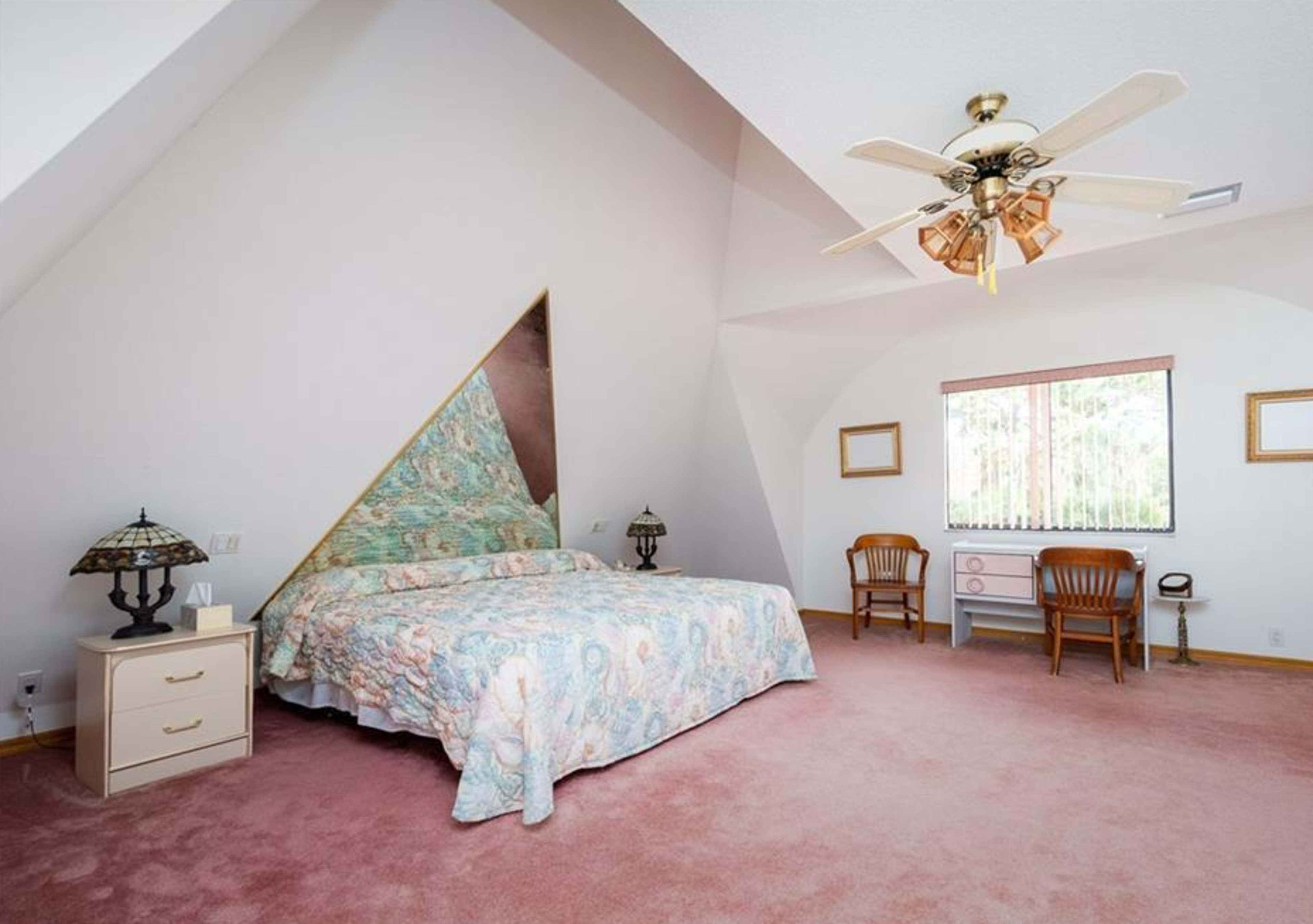 A spacious bedroom features an angled ceiling, a patterned bedspread, wooden furniture, and a window with decorative blinds.