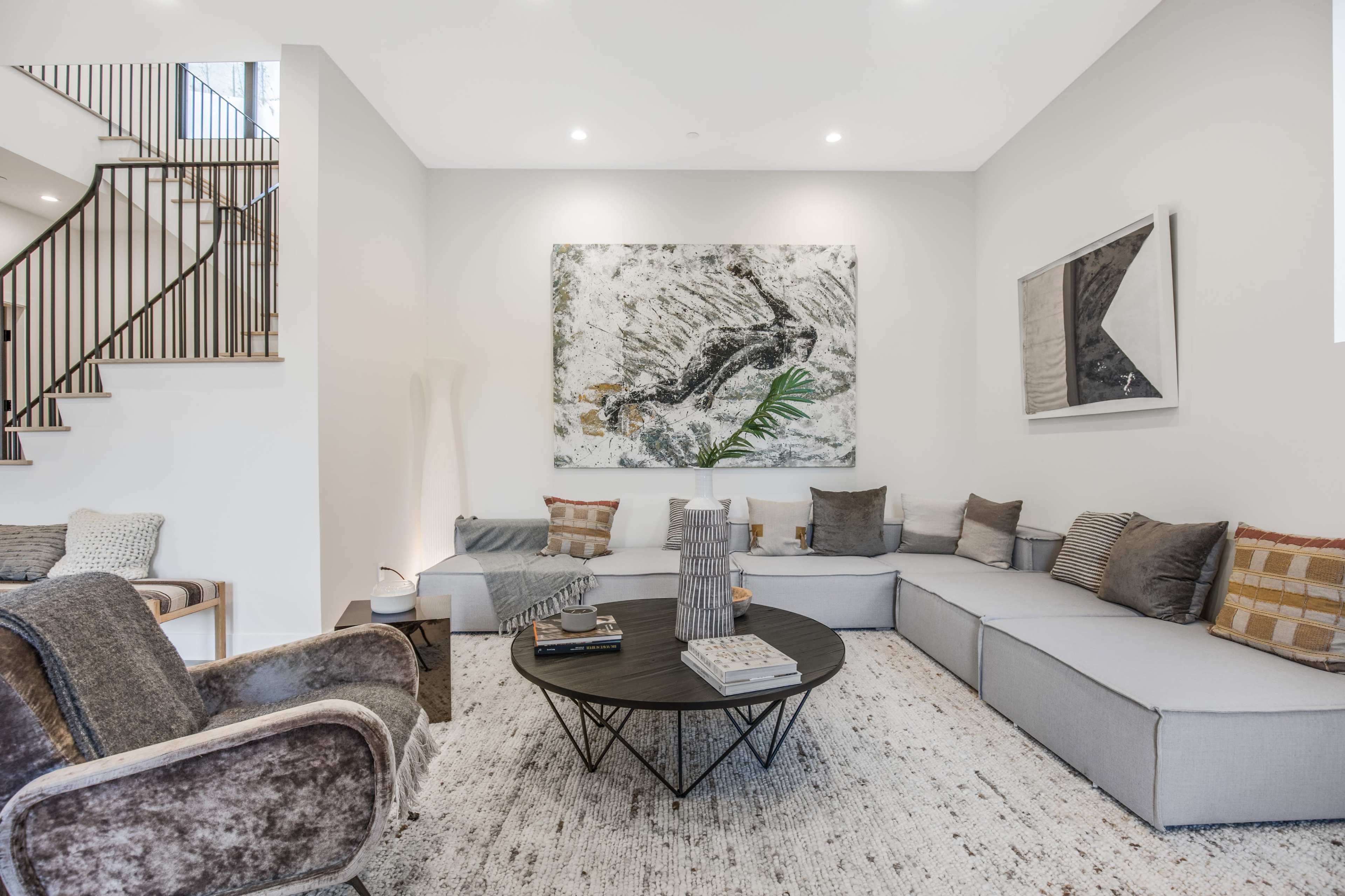 The image shows a modern living room with a large sectional couch, a round coffee table in the center, and a staircase in the background.