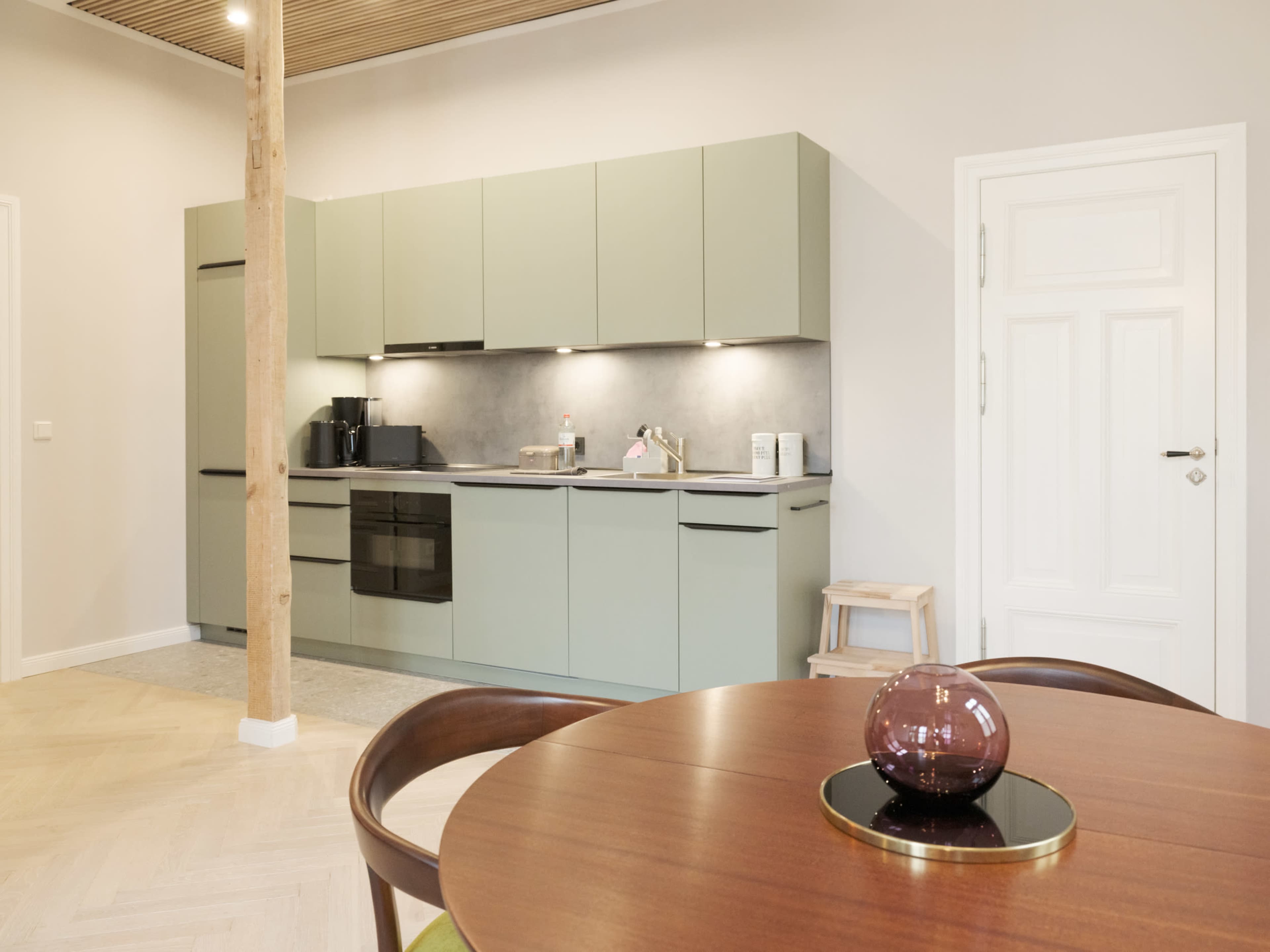 A modern kitchen features green cabinets, a gray countertop, and a wooden table with a decorative glass object.