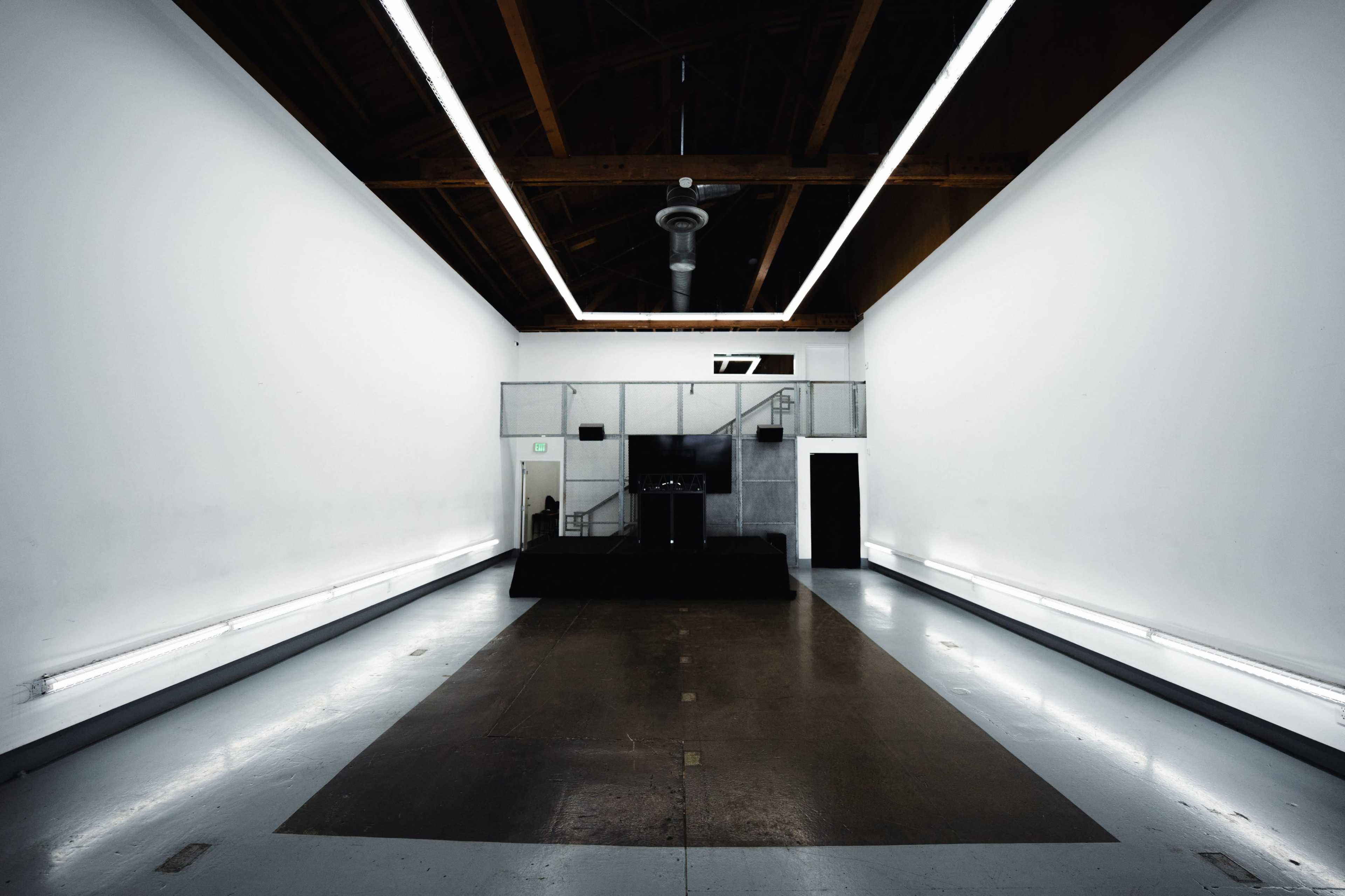 The image shows an empty, spacious interior with stark white walls, a wooden ceiling, and a dark platform at one end.