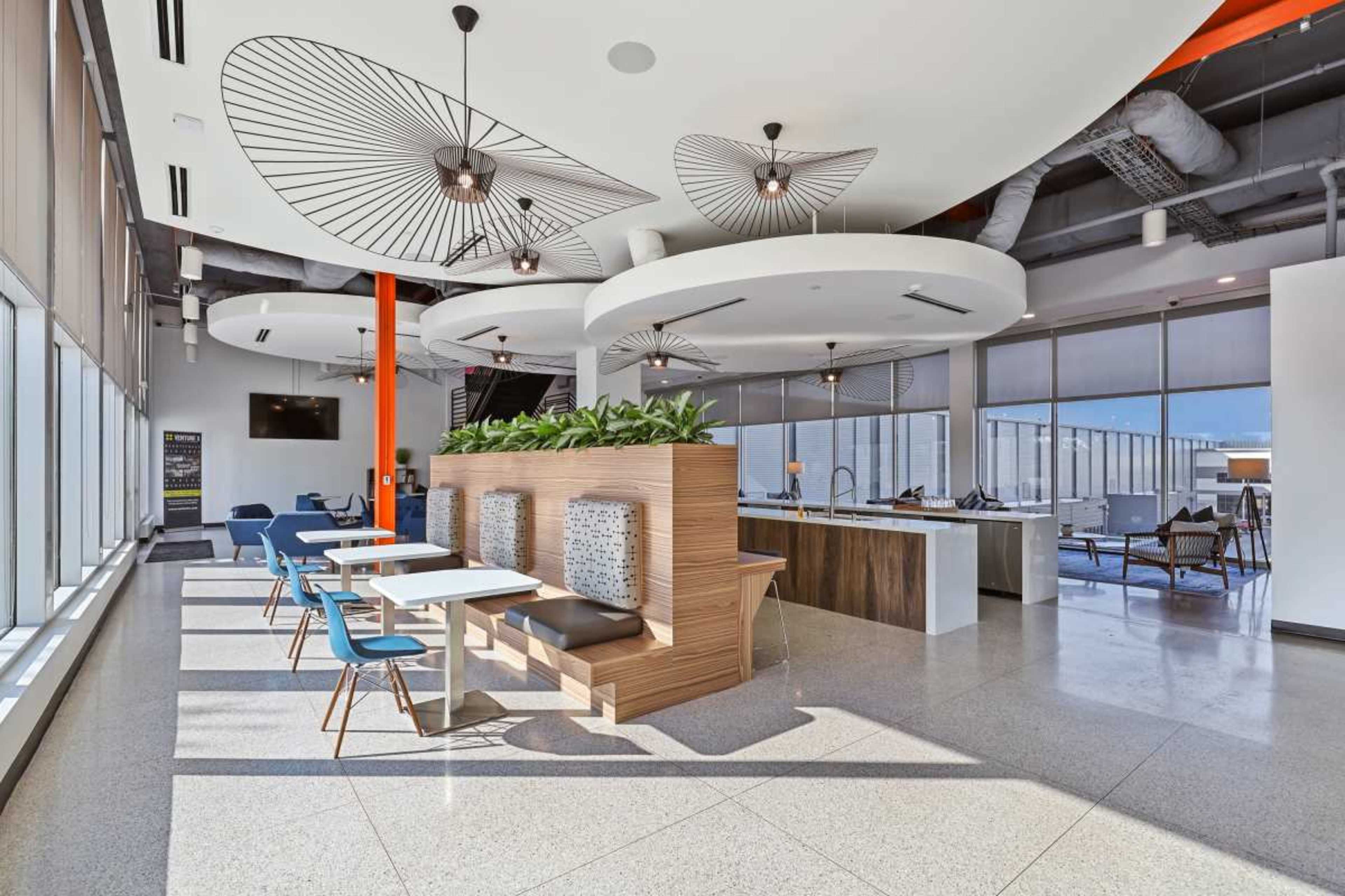 The image shows a modern office space featuring open seating areas, stylish light fixtures, and large windows allowing natural light to fill the room.