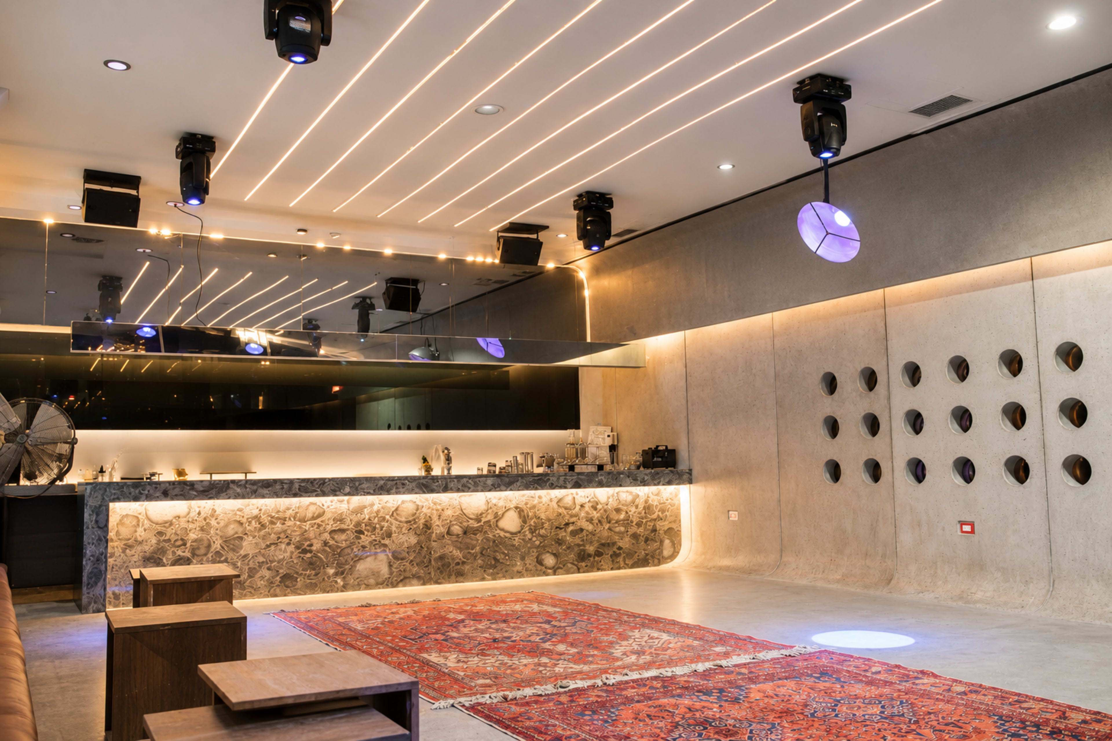 The image shows a modern interior space featuring a bar area with a stone counter, soft lighting, and patterned rugs on a concrete floor.