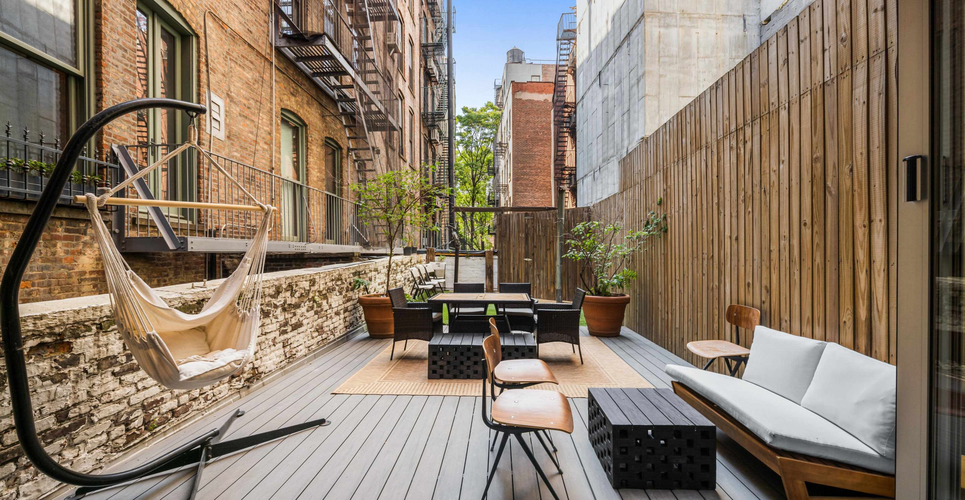A private balcony with wooden decking, featuring a hammock, a seating area, and plants alongside brick and wooden walls.