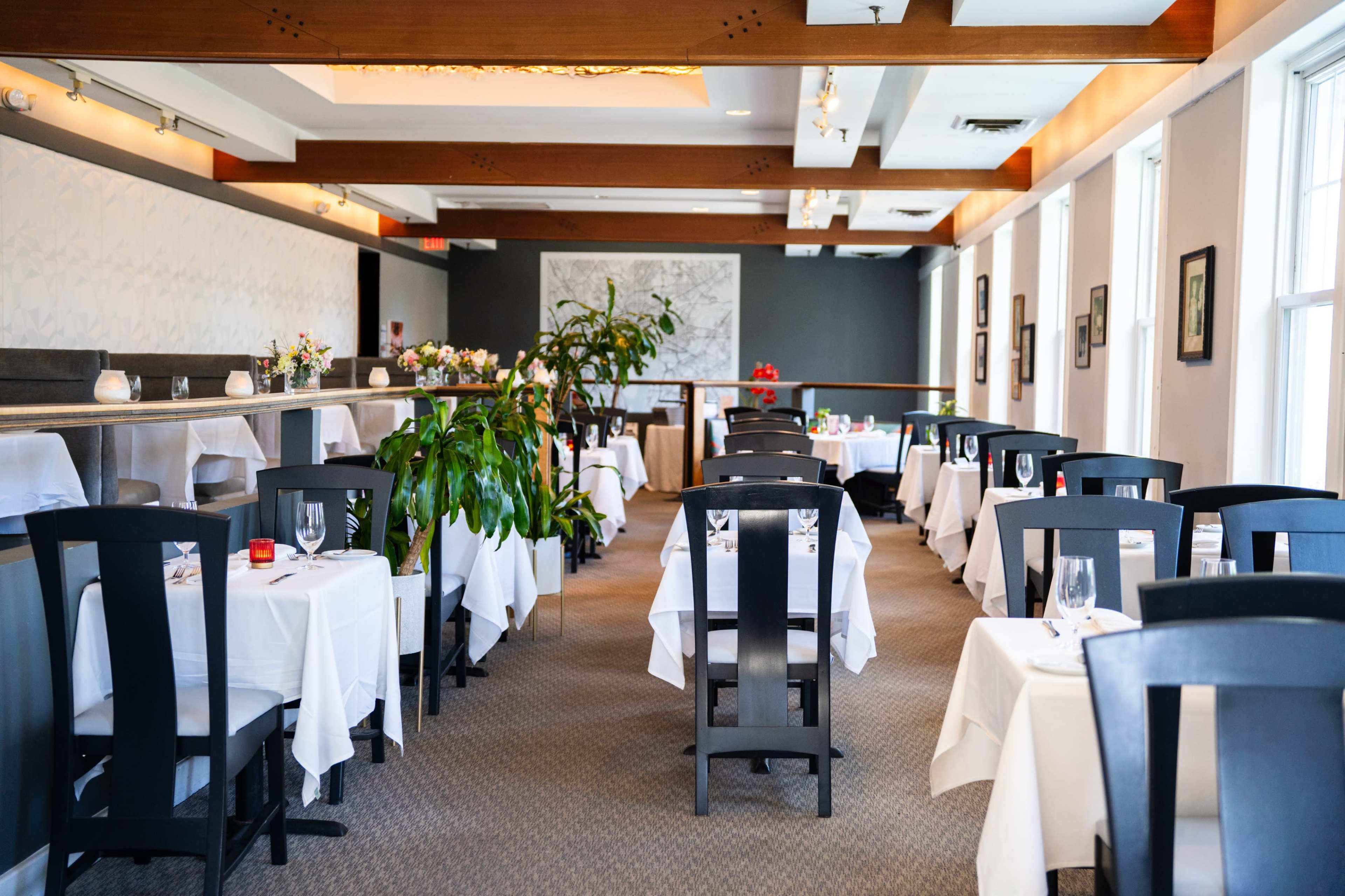 The image shows a restaurant interior with neatly arranged tables covered in white tablecloths and a variety of decorative plants.