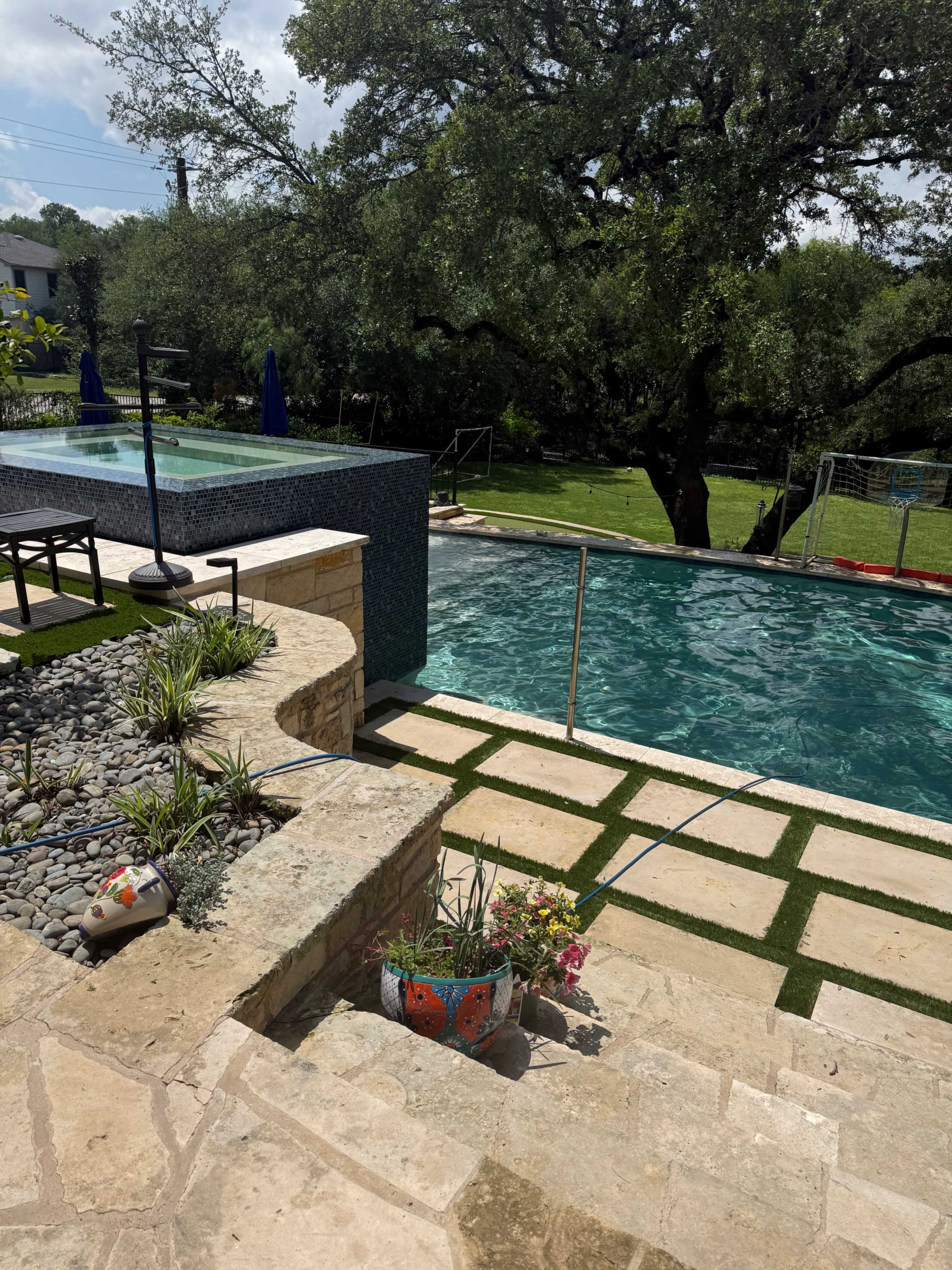 The image shows a modern outdoor swimming pool surrounded by landscaping, with steps leading down from a stone patio to the pool area.