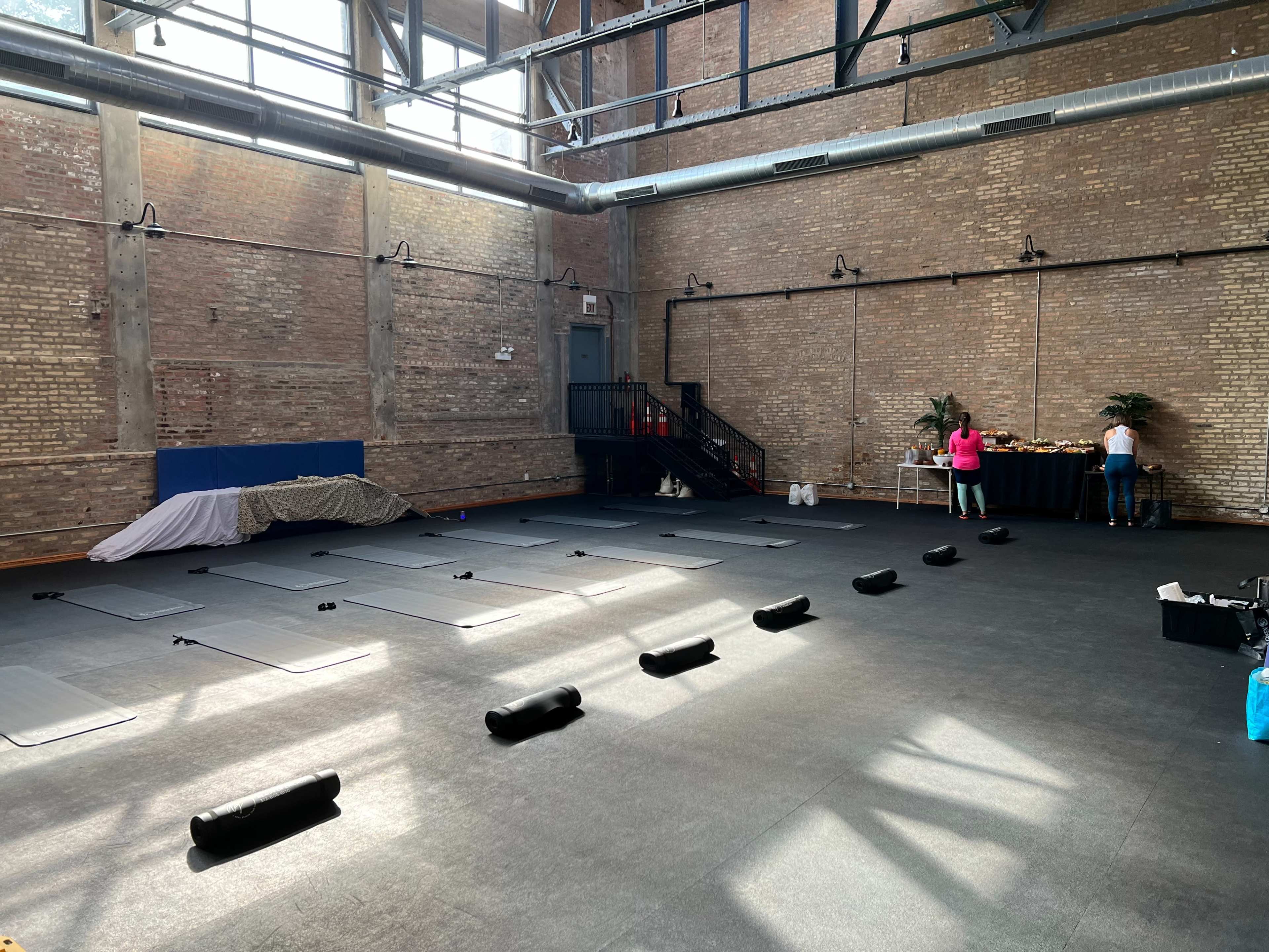 The image shows a spacious, empty studio with yoga mats and props arranged on the floor, and a table with refreshments in the corner.