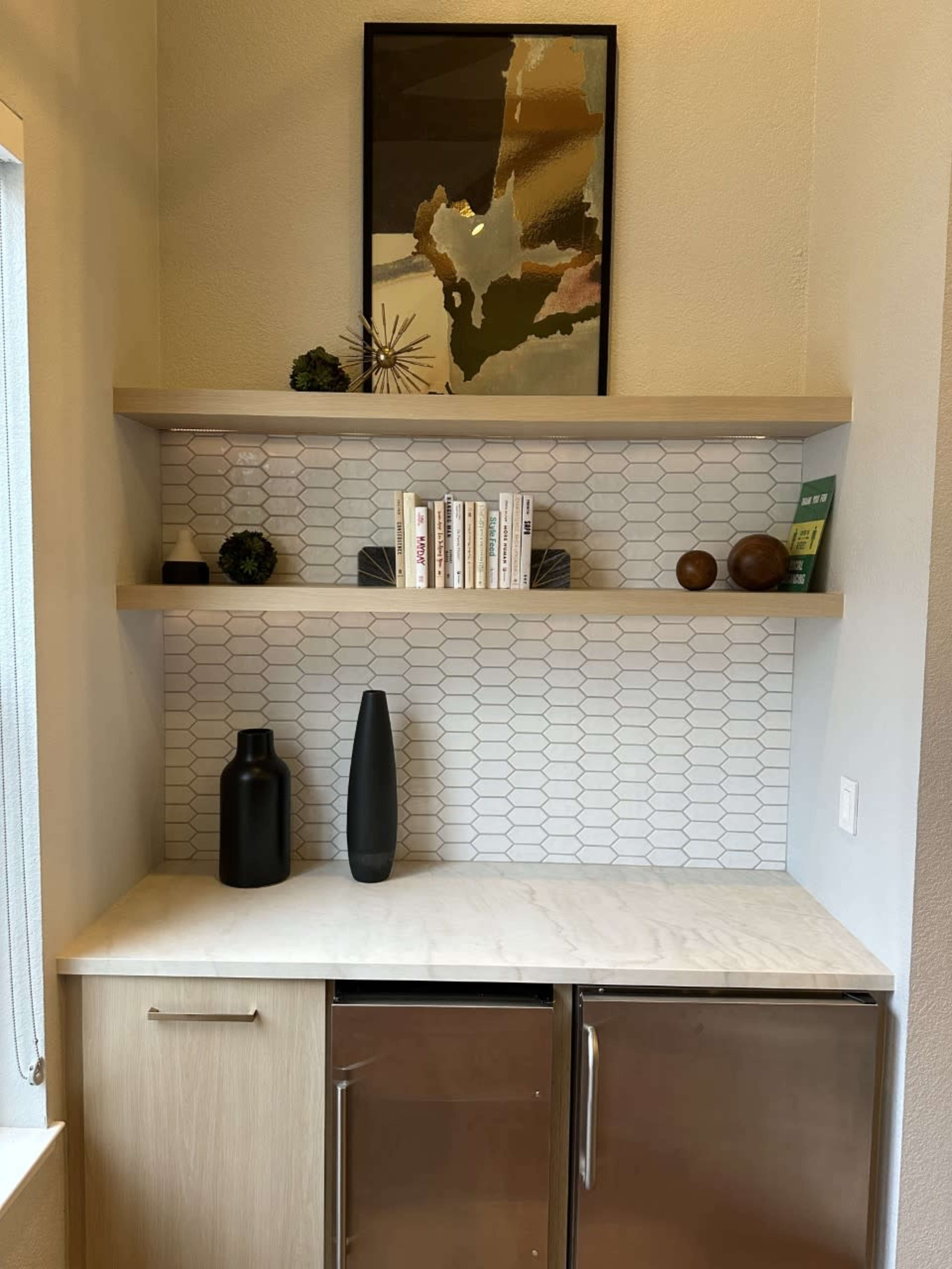 The image shows a modern kitchenette corner with a marble countertop, metallic refrigerator, open shelves displaying books and decor, and a textured tiled backsplash.