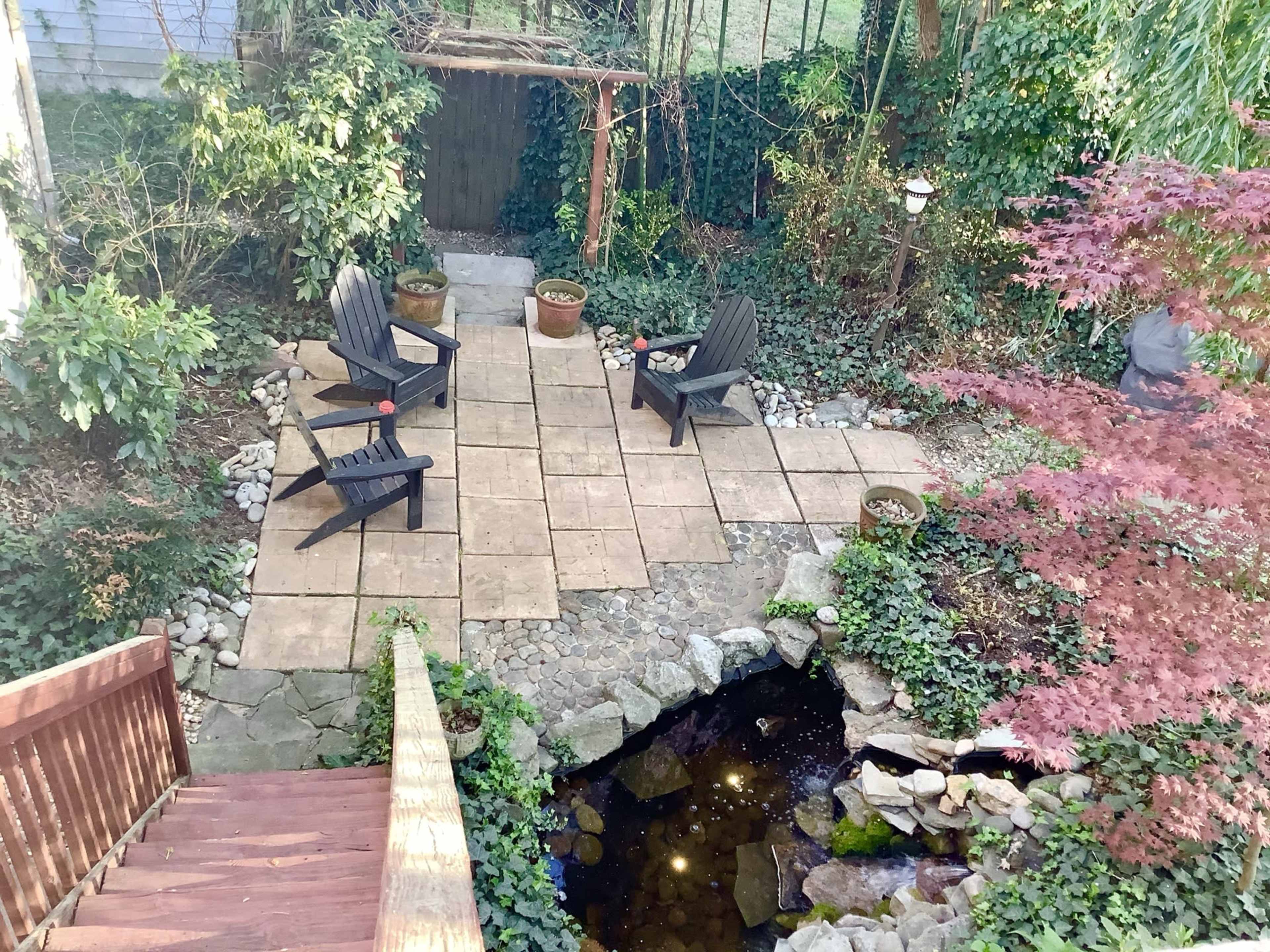 A landscaped backyard with a stone patio, two black Adirondack chairs, potted plants, and a small pond surrounded by greenery.