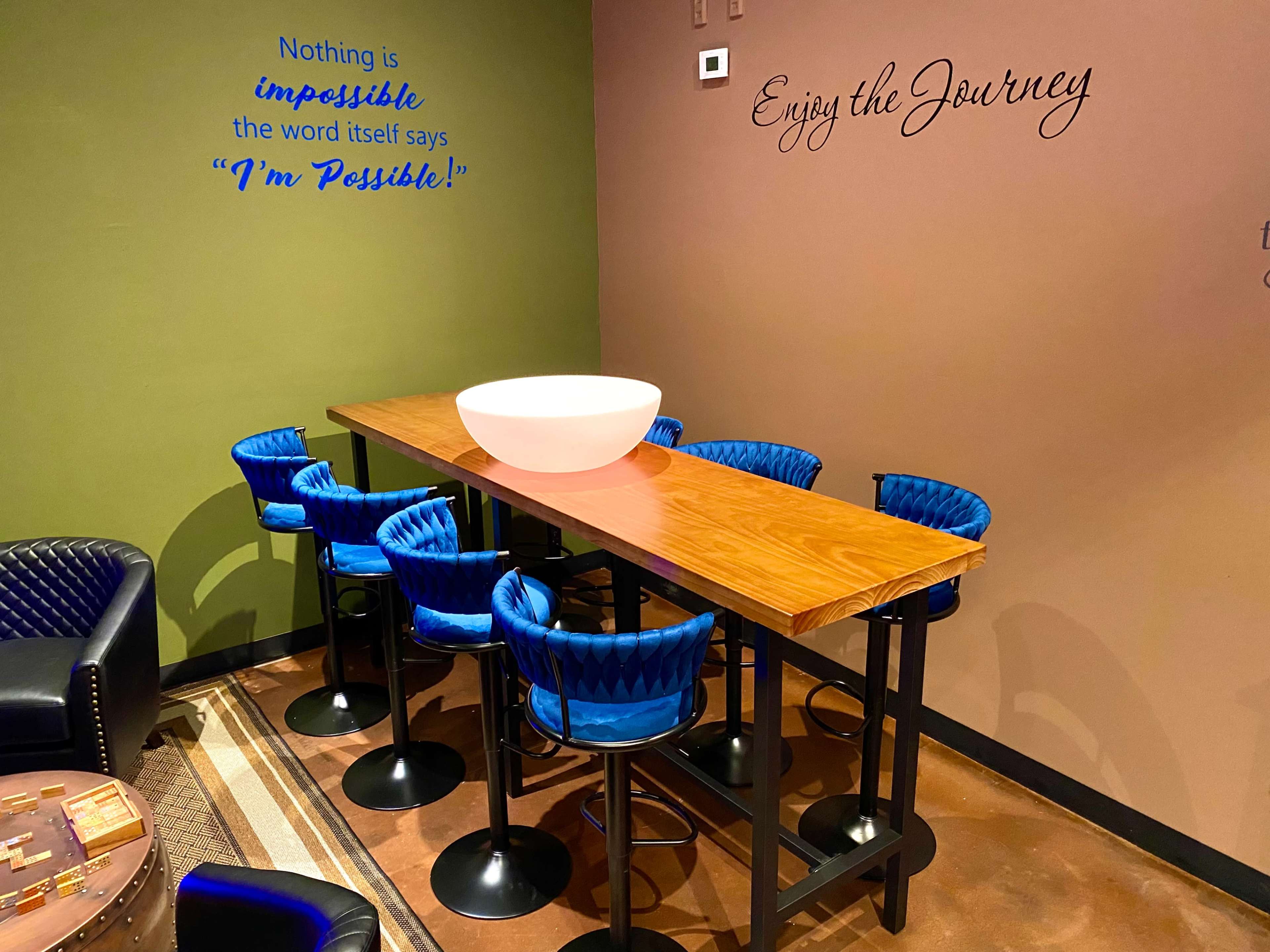 The image shows a modern seating area featuring a long wooden table with a bowl centerpiece and six blue bar stools, set against a backdrop of motivational quotes on the walls.