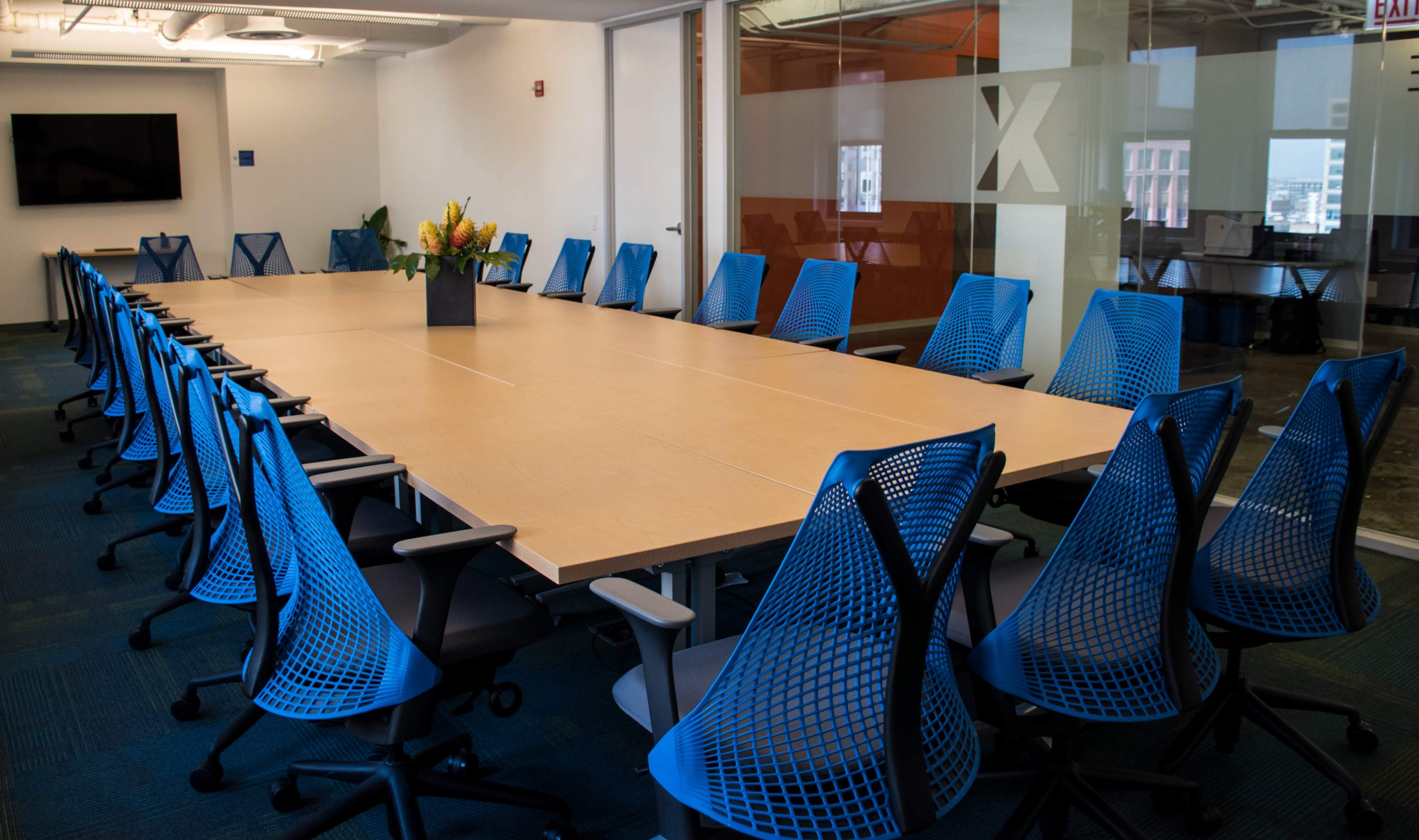 A large conference room features a long wooden table surrounded by blue ergonomic chairs and a central flower arrangement.