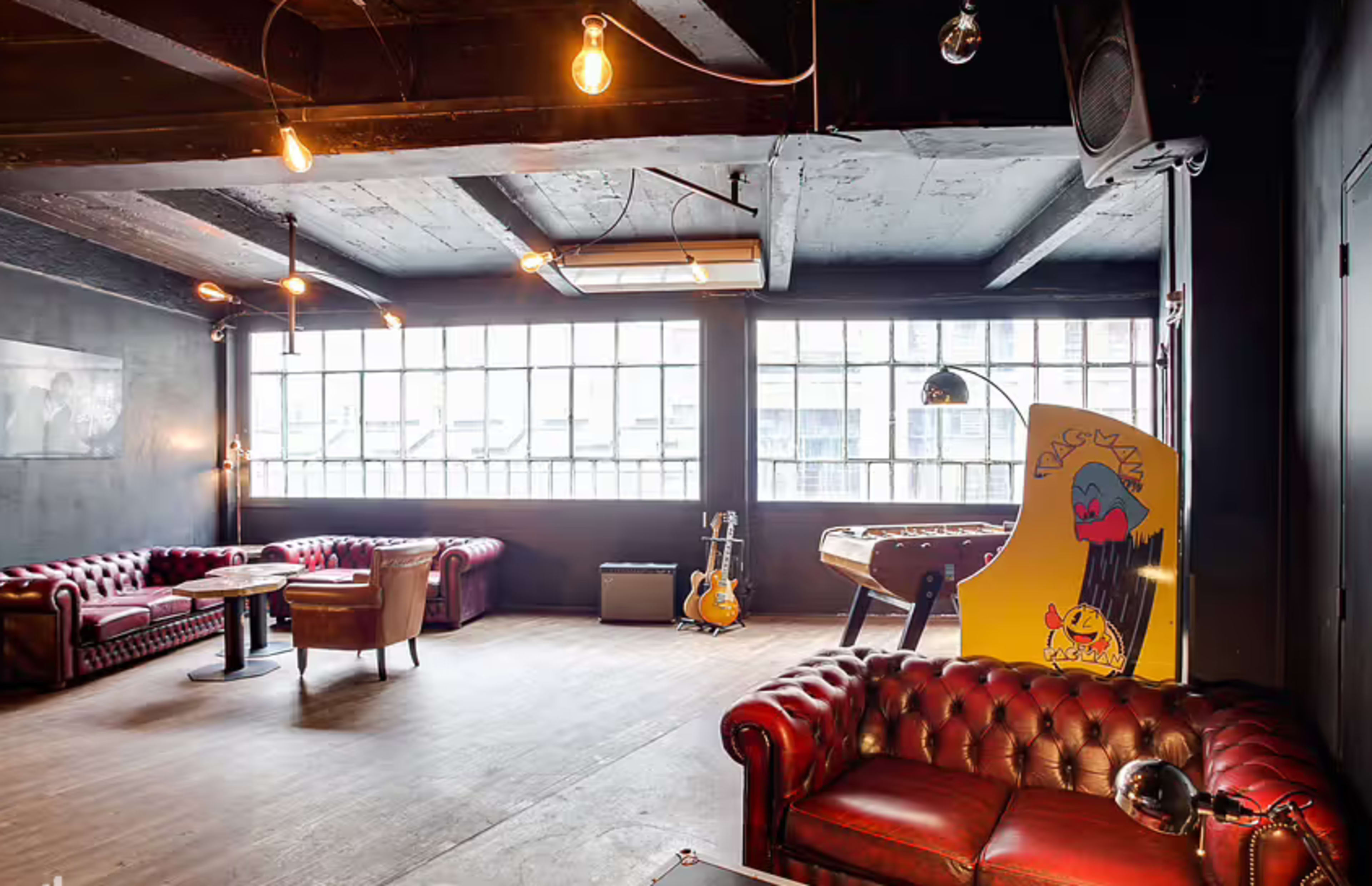 The image shows a spacious lounge area featuring red leather couches, a wooden floor, and large windows, with a vintage arcade game and musical instruments in the background.