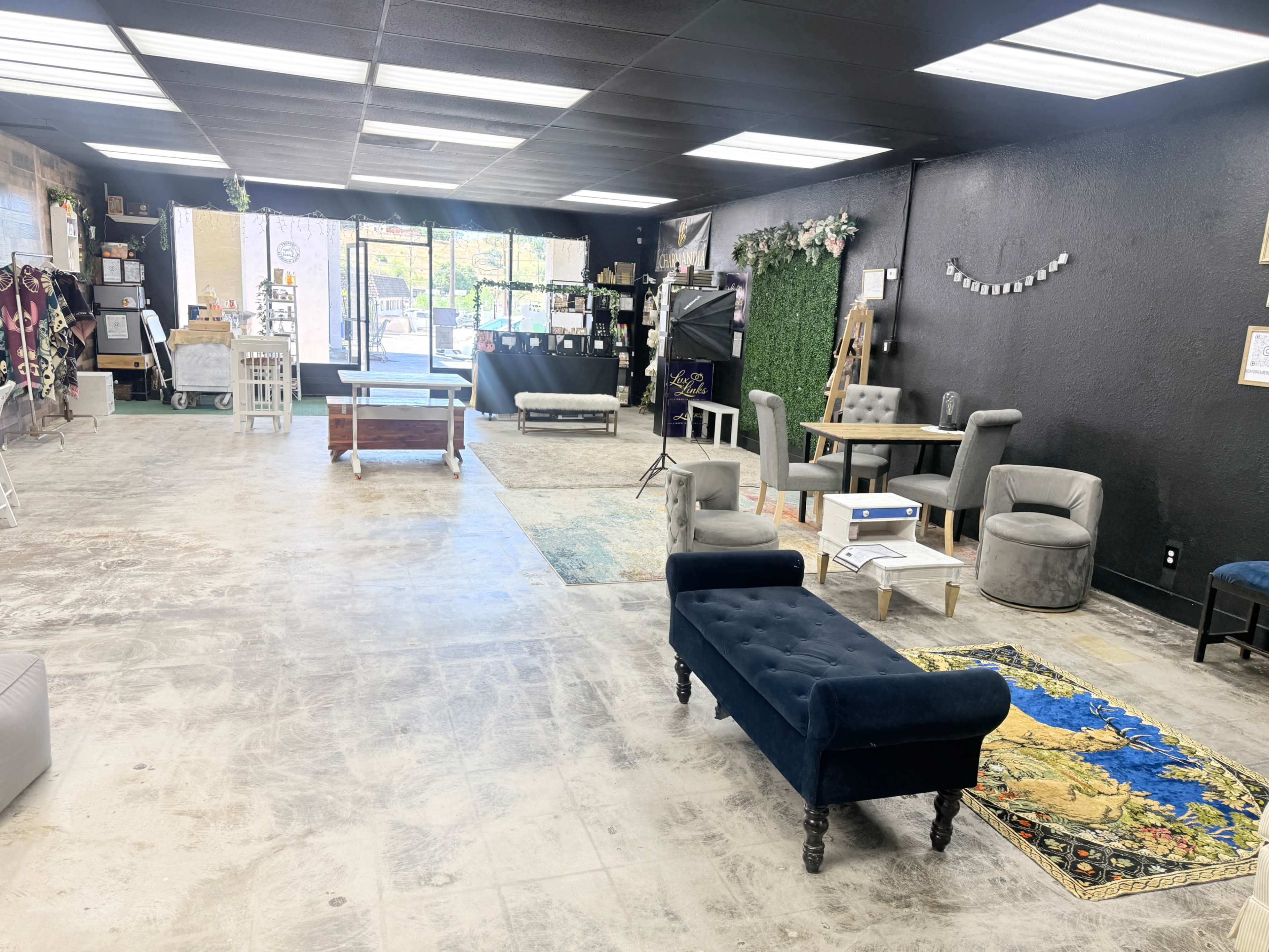 The image shows an empty commercial space with various seating arrangements, a patterned rug, and large windows at the front.