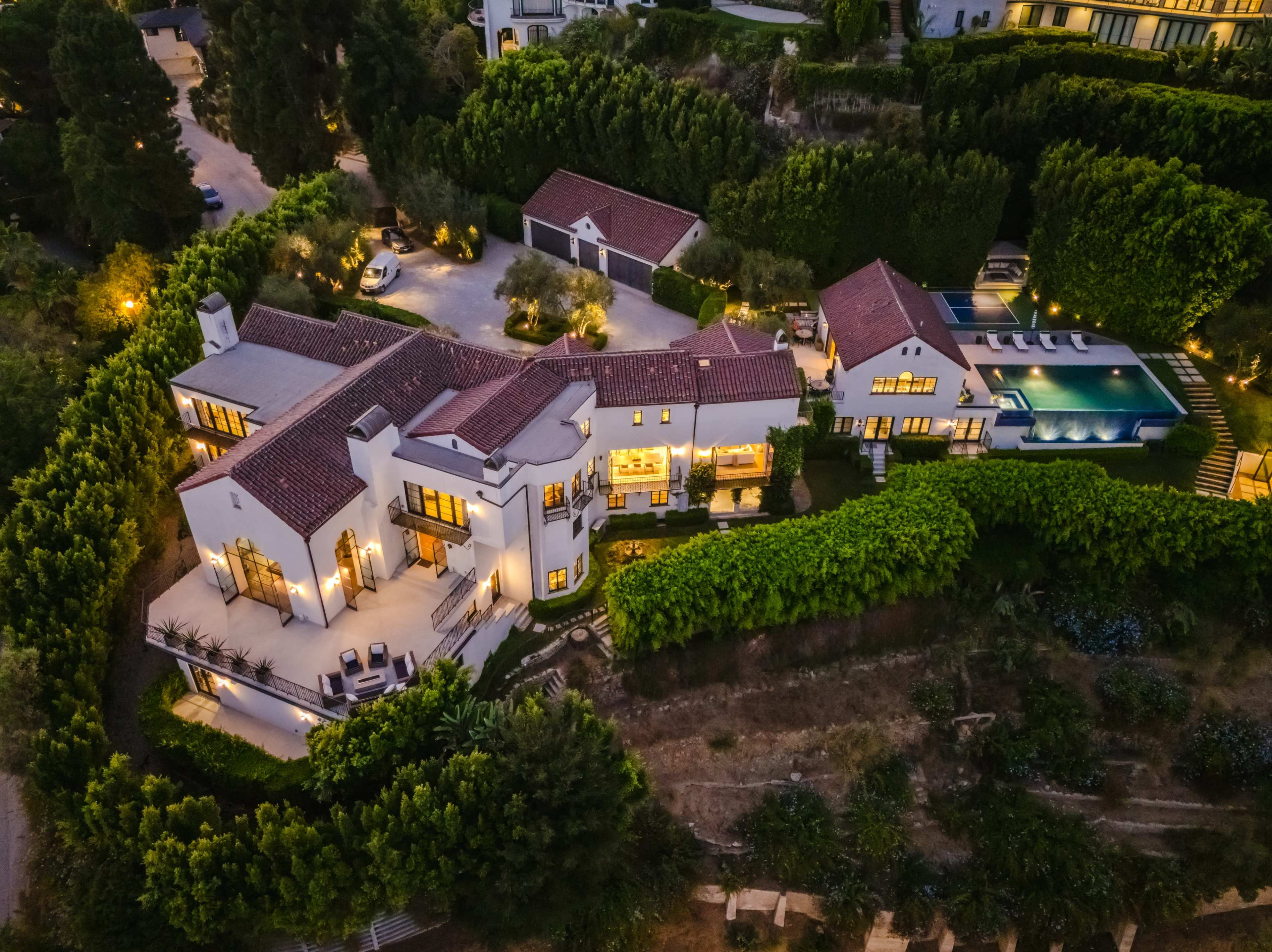The image shows a large, multi-structure residential estate with a swimming pool, surrounded by lush greenery and illuminated pathways.