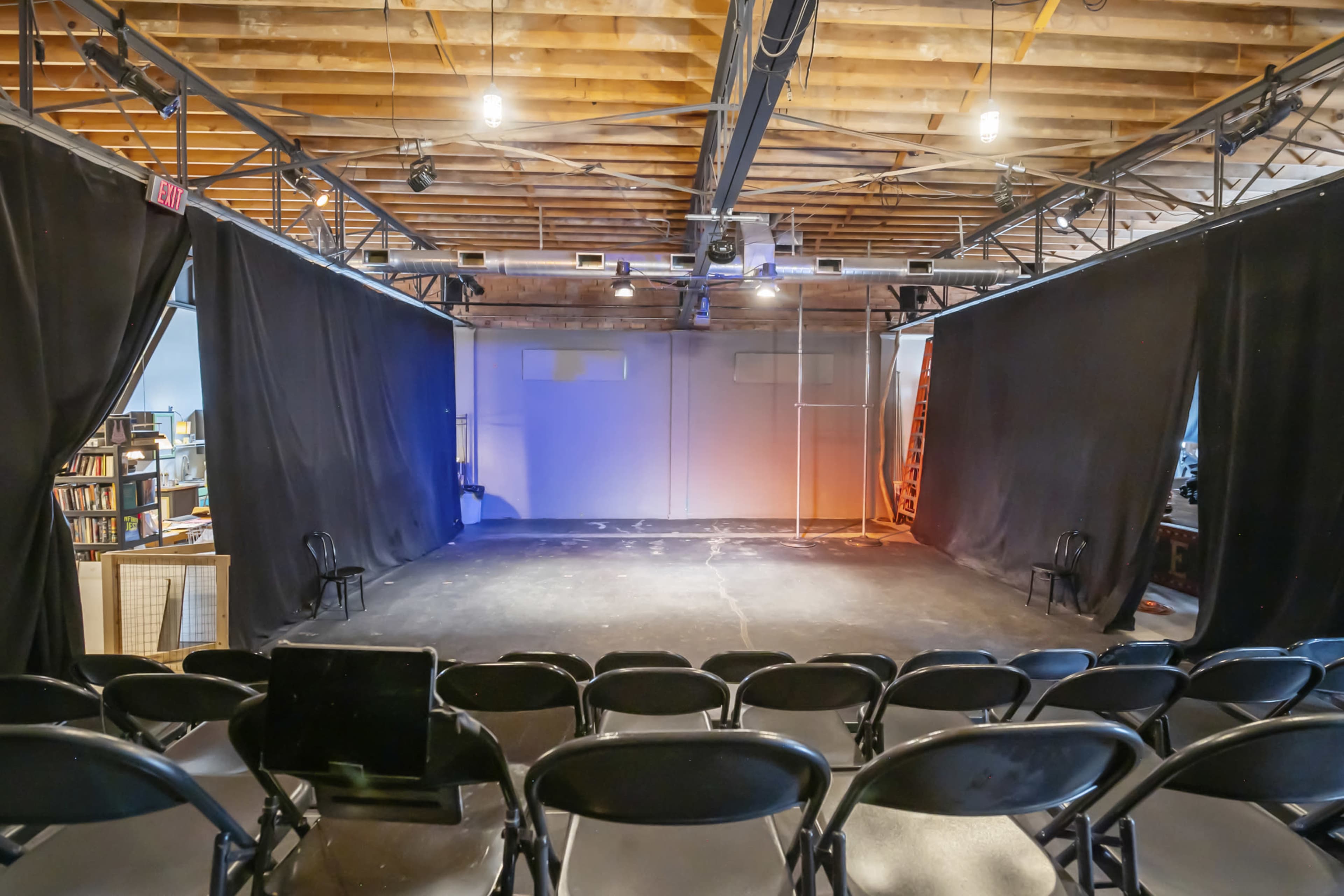 A blank stage in a theater with black curtains, rows of chairs facing the stage, and spotlights overhead.