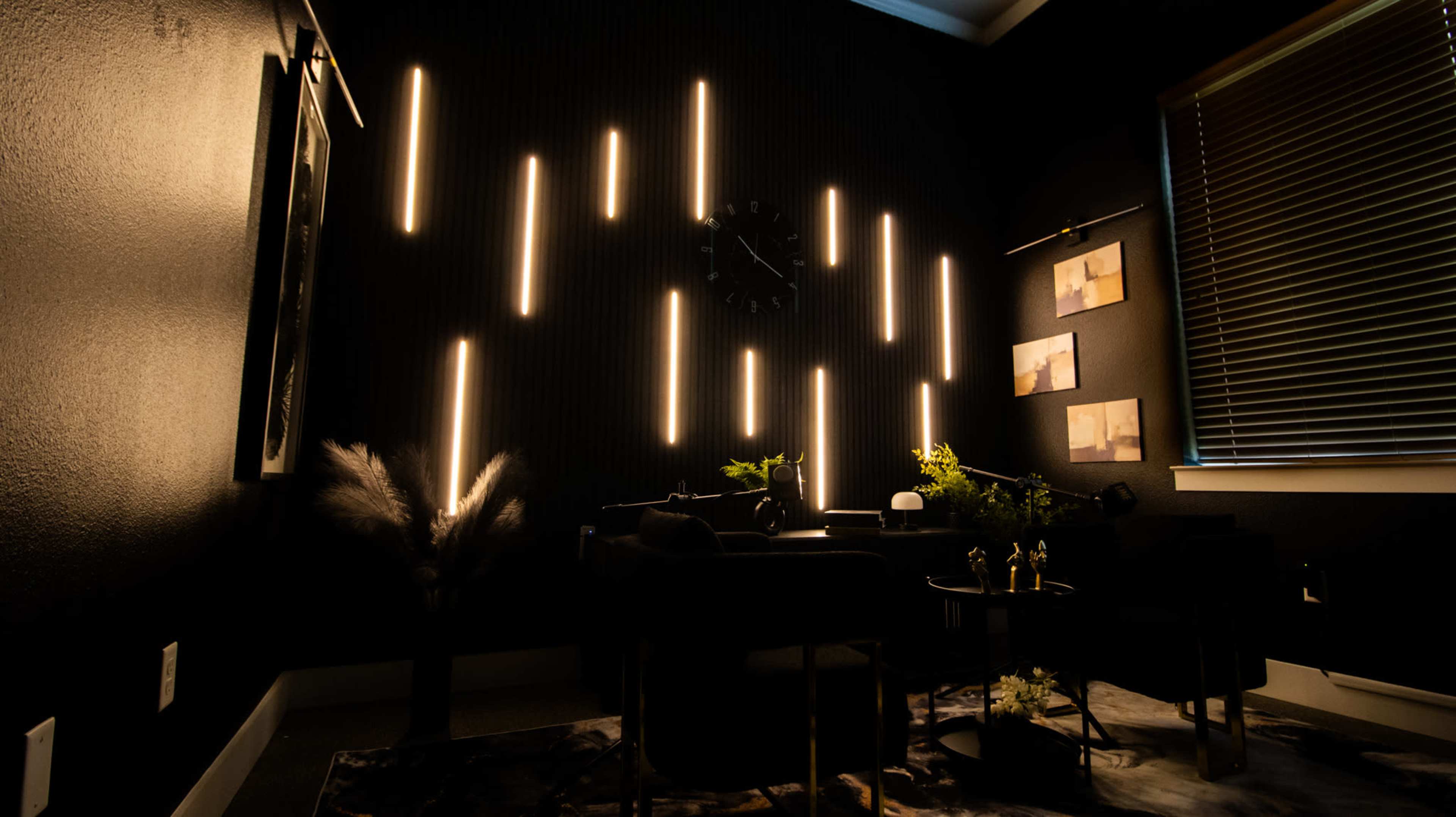 The image shows a dimly lit room with vertical light strips on a dark wall, a clock, and decorative plants in a modern interior setting.