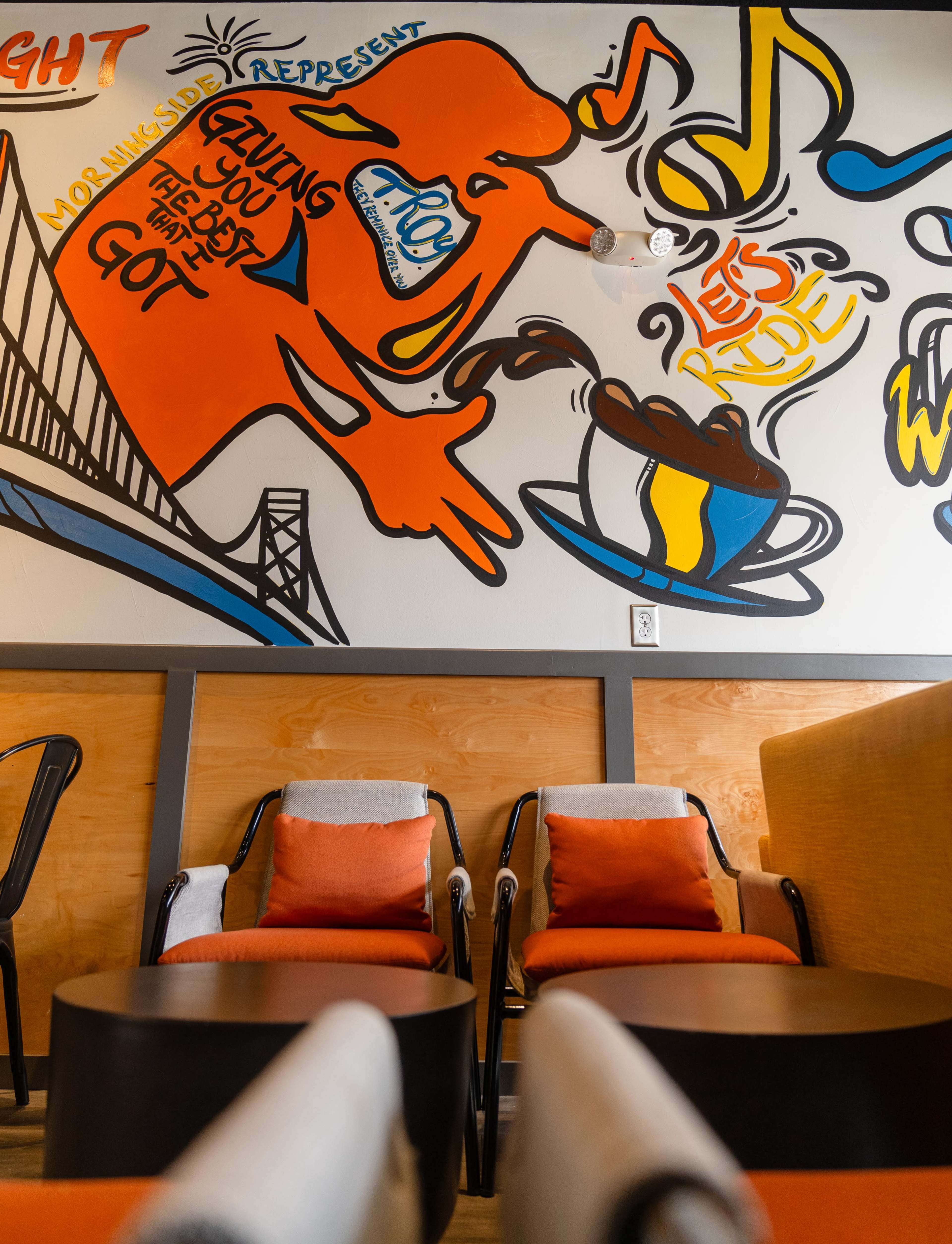 A colorful mural featuring a stylized figure and musical notes on a wall in a coffee shop, with two chairs and a table in the foreground.