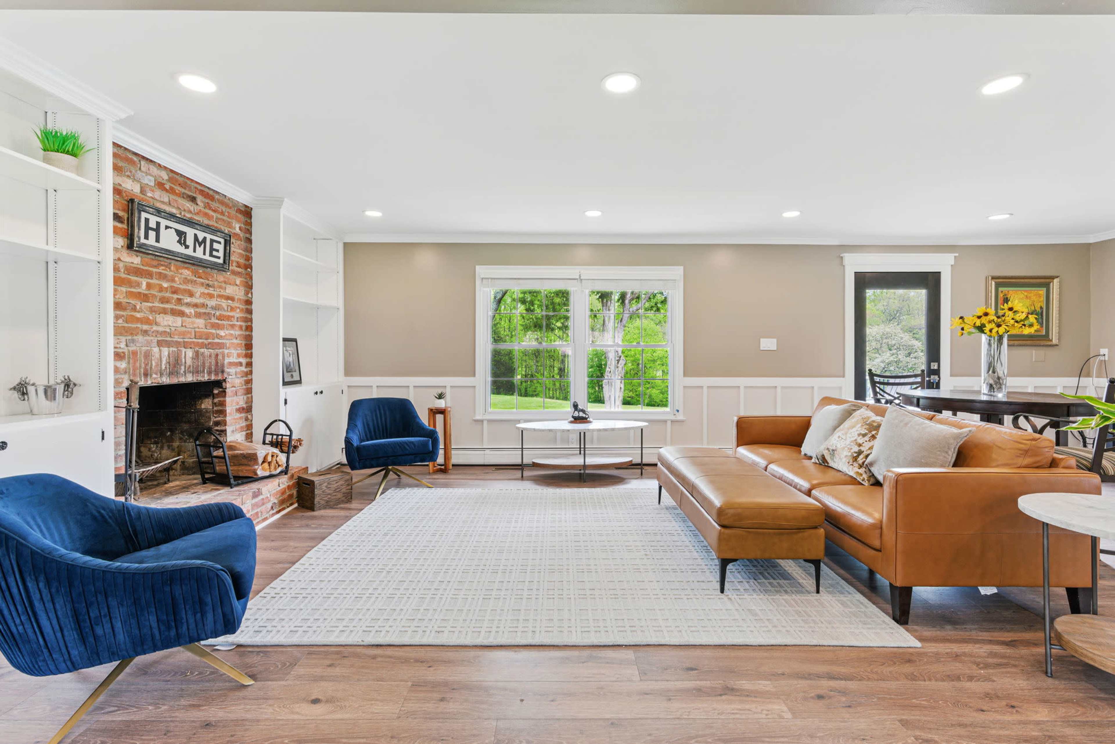 The image shows a modern living room featuring a brown leather sofa, blue armchairs, a white area rug, and a brick fireplace, with large windows allowing natural light to fill the space.