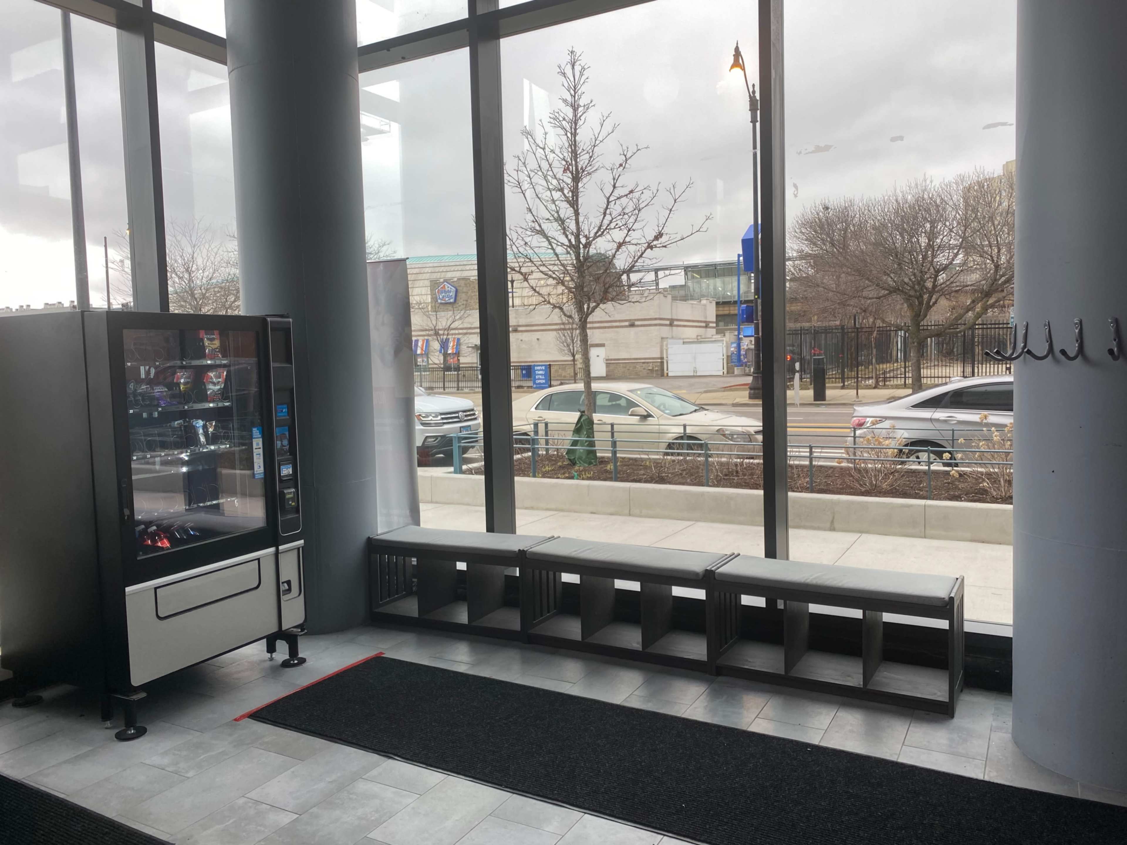 A vending machine next to a bench, with large windows overlooking a street and parked cars outside.