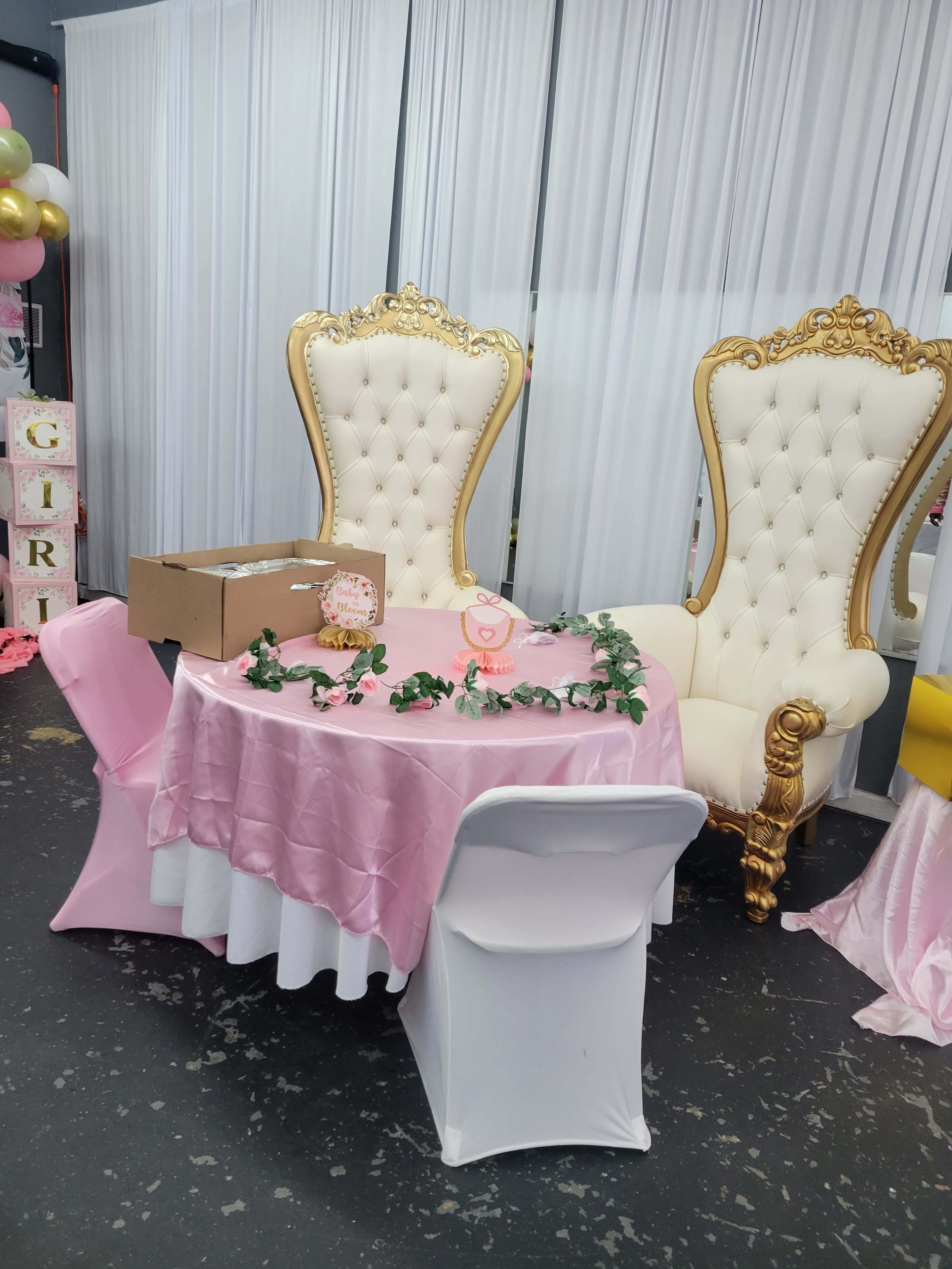 Two ornate golden chairs flanking a small pink table decorated with greenery, situated in an event space with white drapery and balloons.