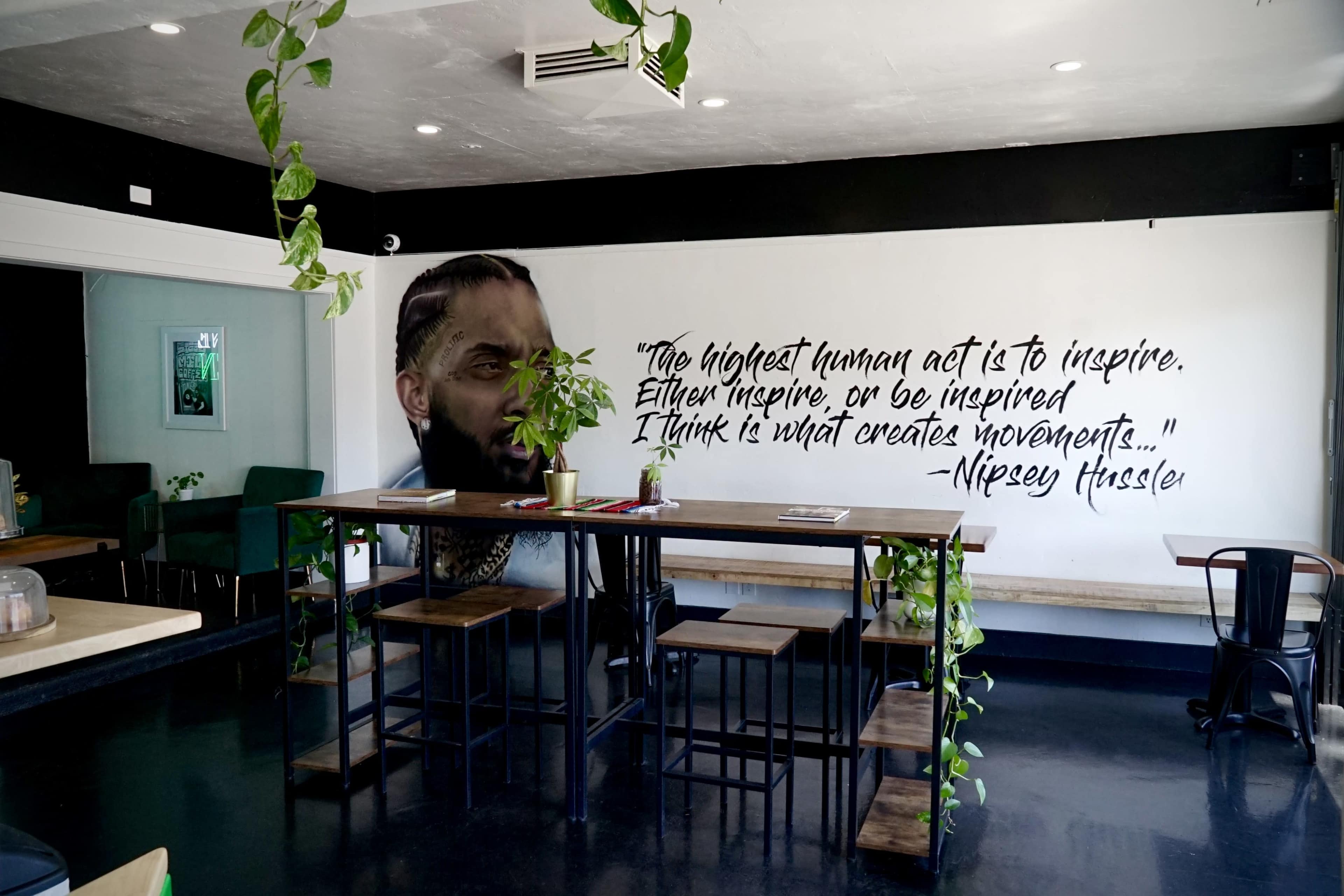 A modern café interior featuring a large mural of a man's face alongside a quote about inspiration, with a wooden table and plants in the foreground.