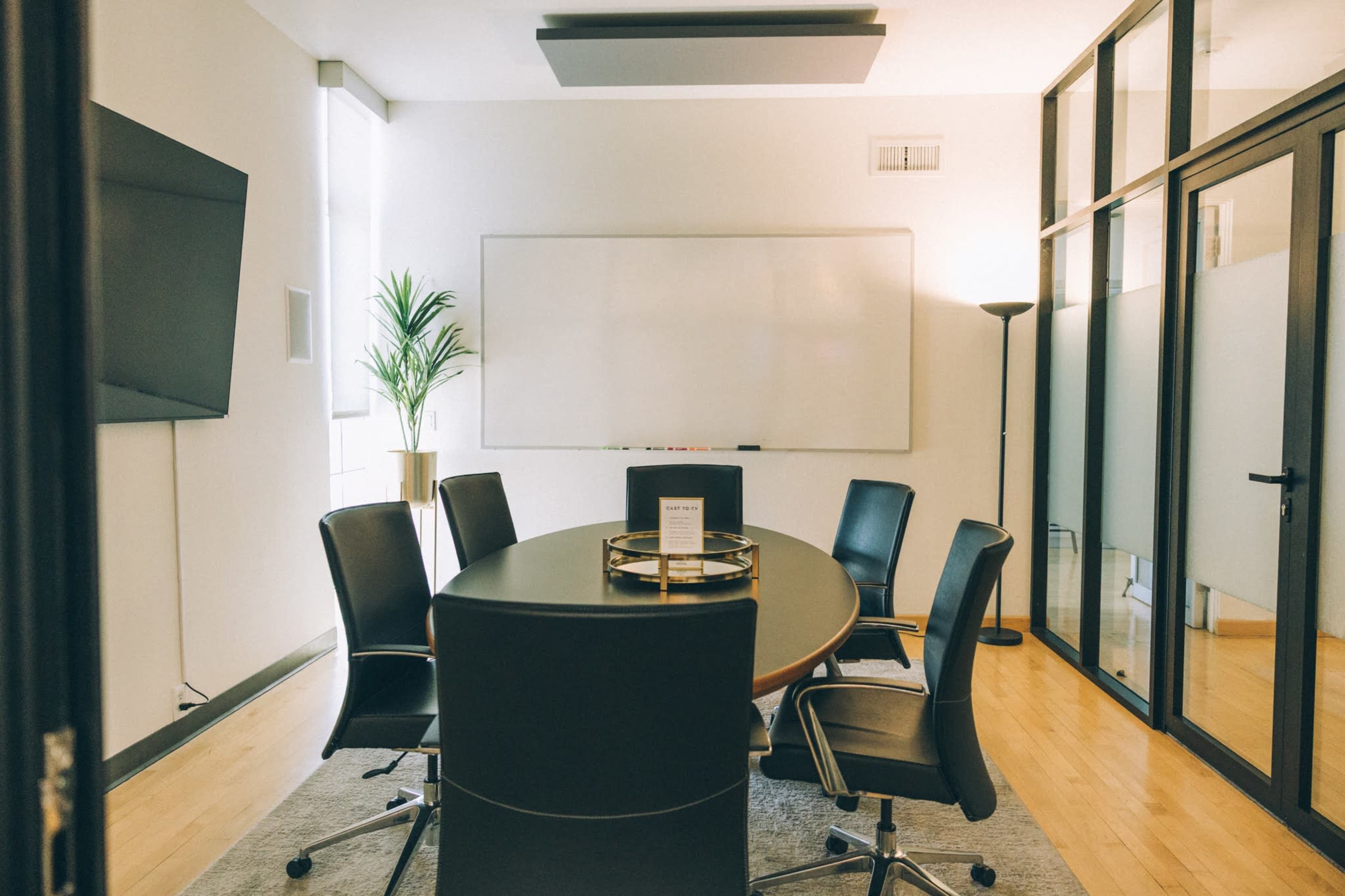 A modern conference room features a large oval table surrounded by black chairs, with a whiteboard and a wall-mounted TV in the background.