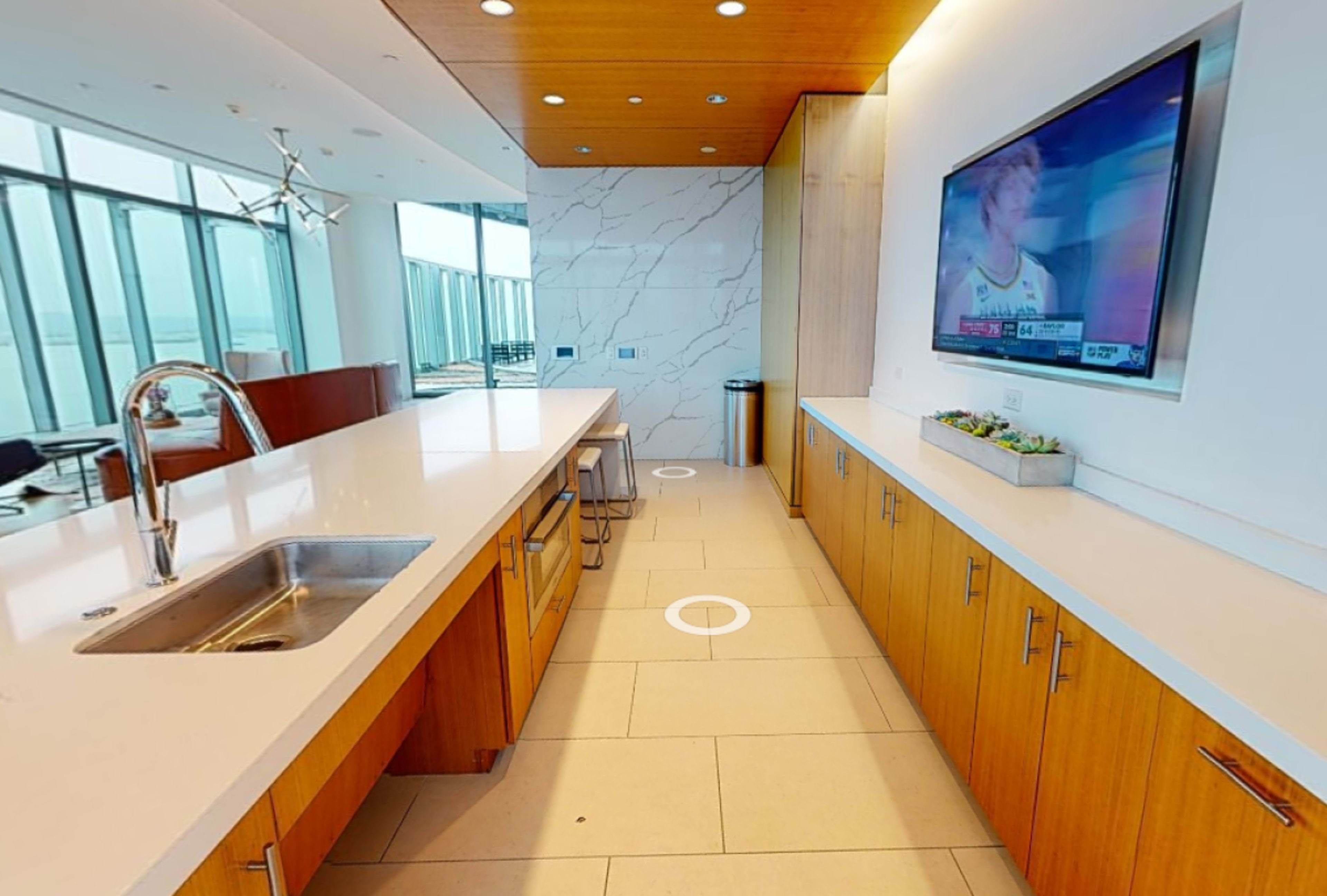 A modern kitchen area features a long countertop with a sink, bar stools, and a television mounted on the wall.