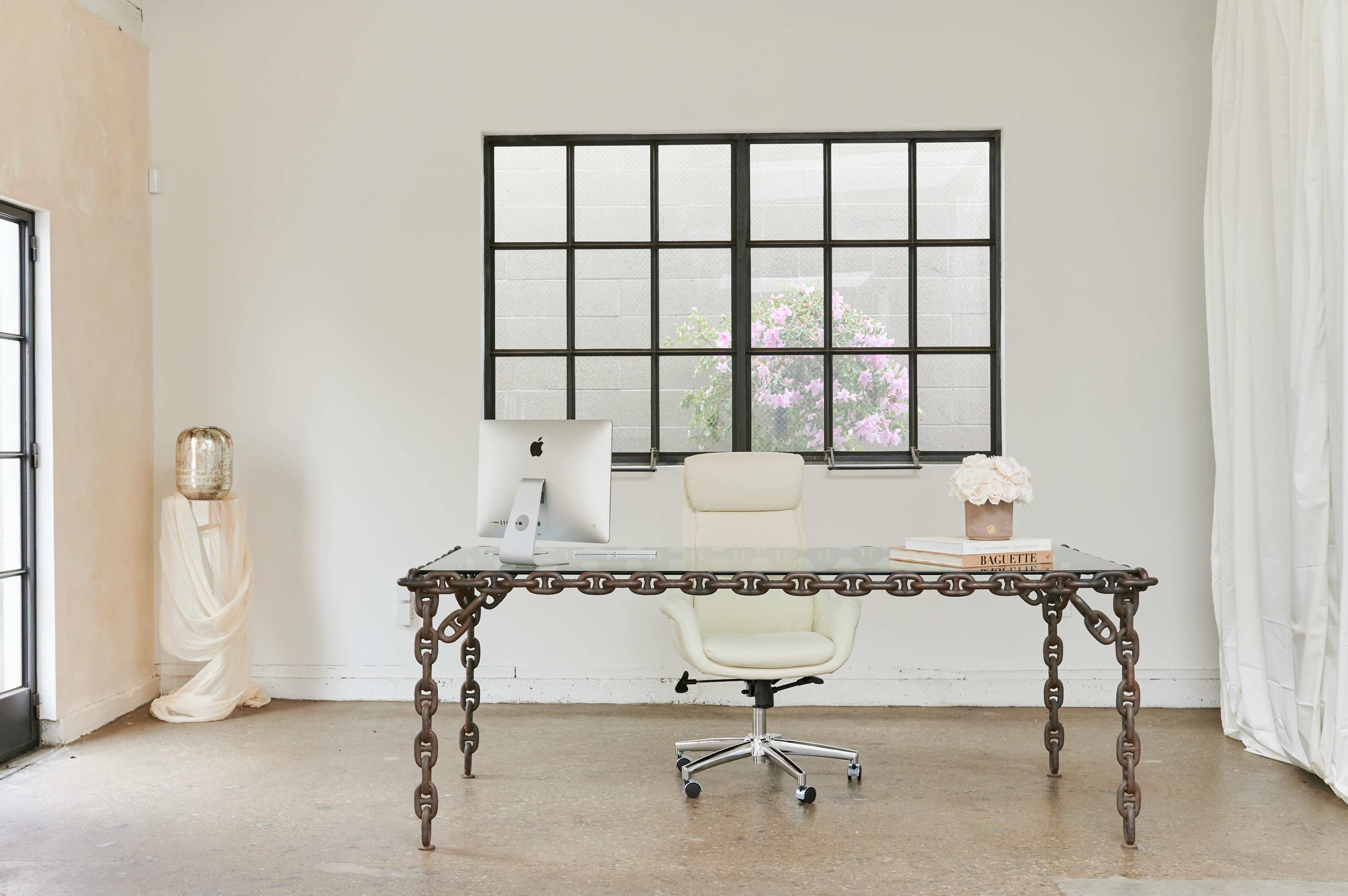 A modern office space features a glass desk with chain-link legs, a computer, and a chair, with a window showing flowering plants outside.