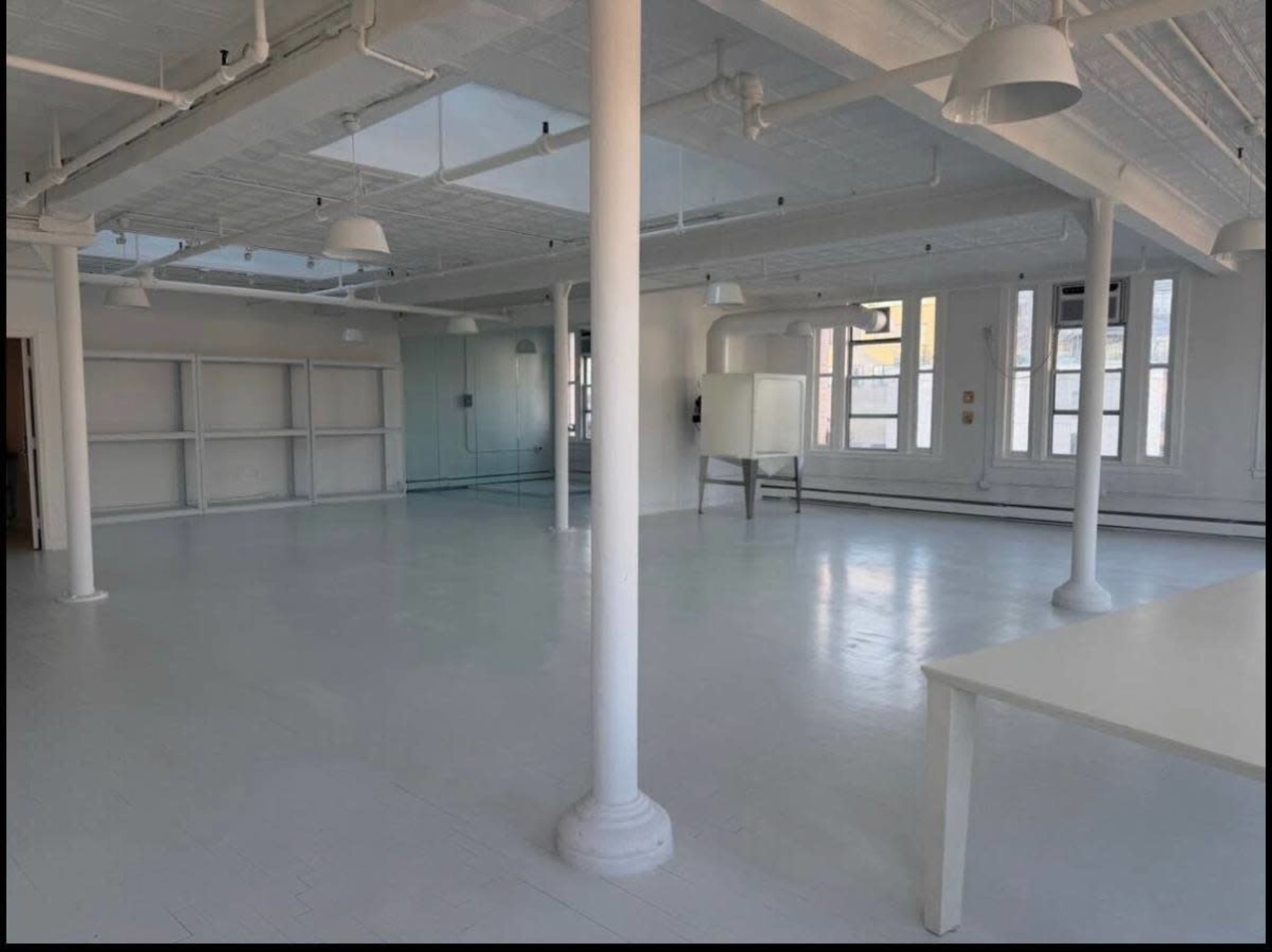 The image shows a spacious, empty room with high ceilings, white walls, and several columns, featuring large windows that allow natural light to enter.