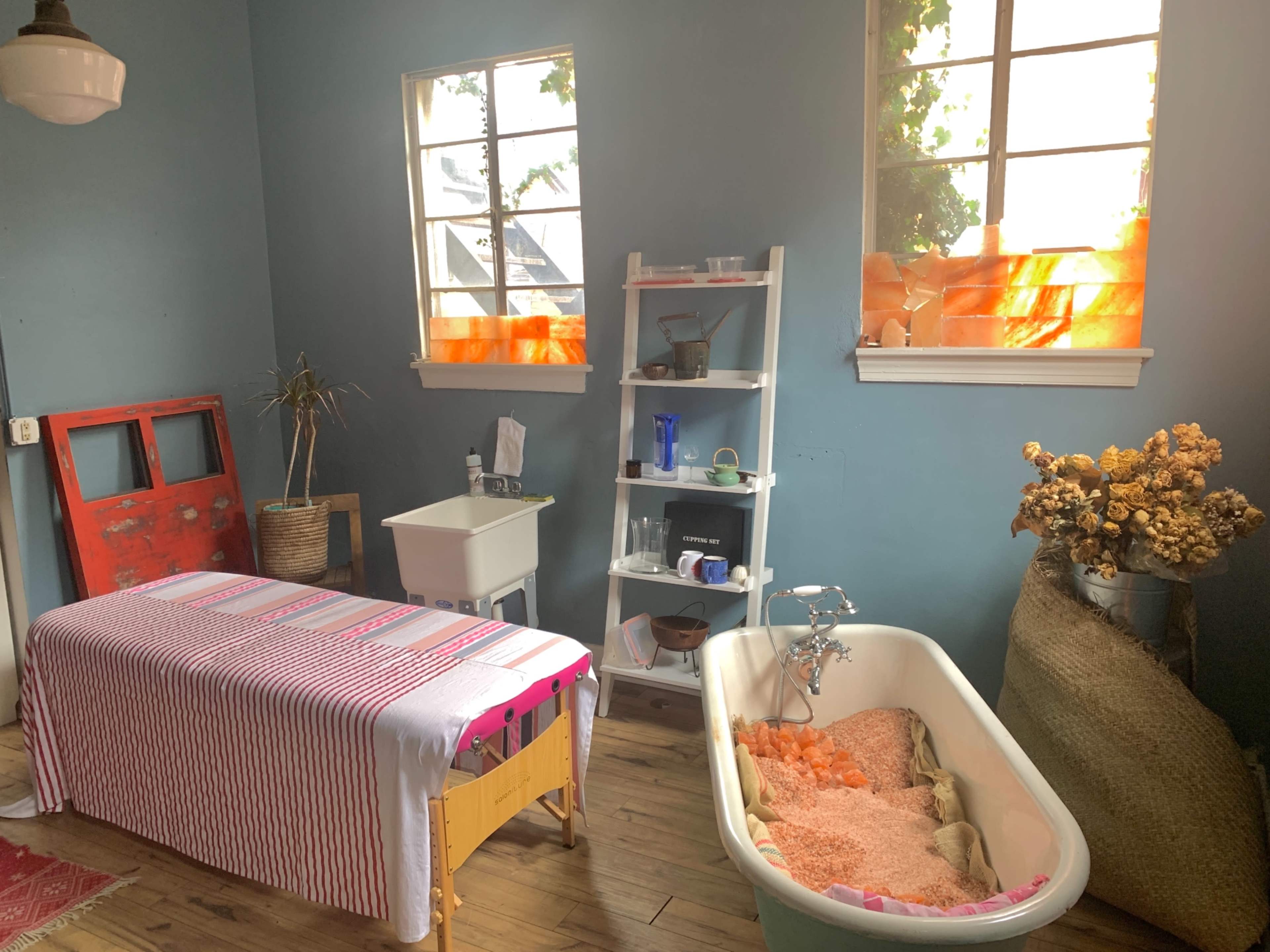 A simple room with a massage table, a vintage bathtub filled with orange bath salts, a white shelf with various items, and two windows with orange curtains.