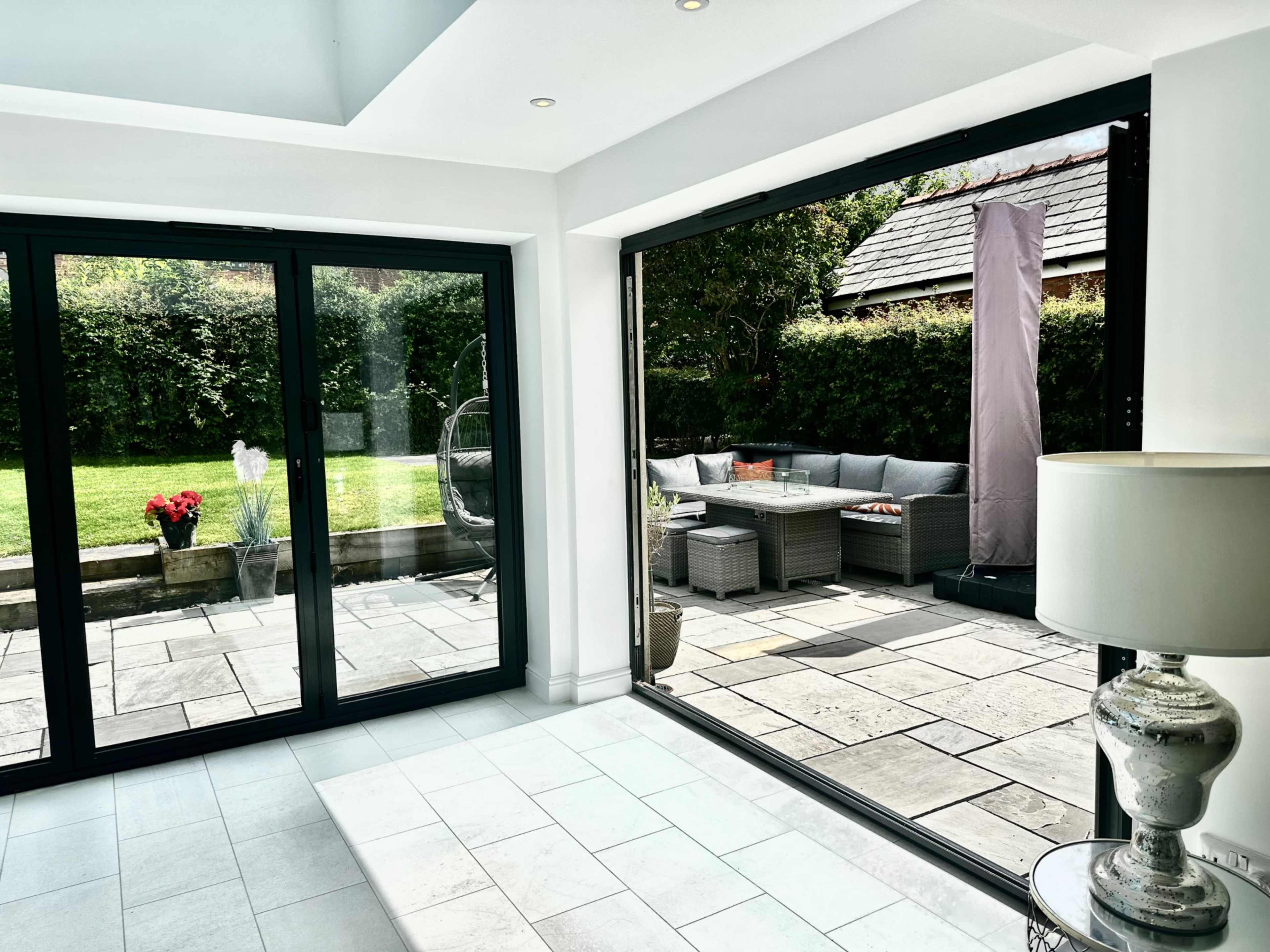 The image shows a modern room with large glass doors opening to a patio area featuring outdoor furniture and a well-maintained garden.