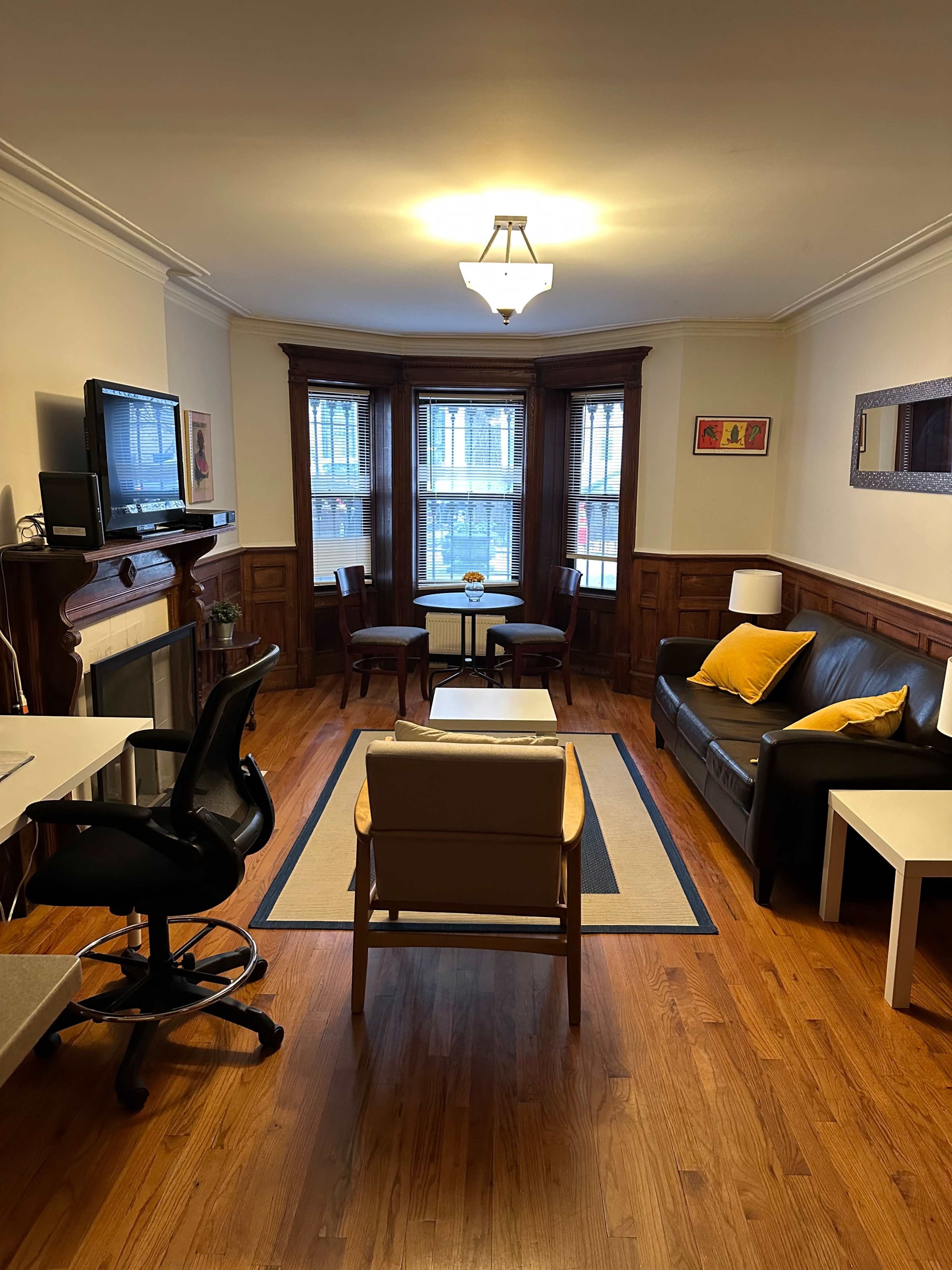 A cozy living room featuring a black sofa, a wooden coffee table, two chairs, a desk with a rolling chair, and a television mounted on a wall.
