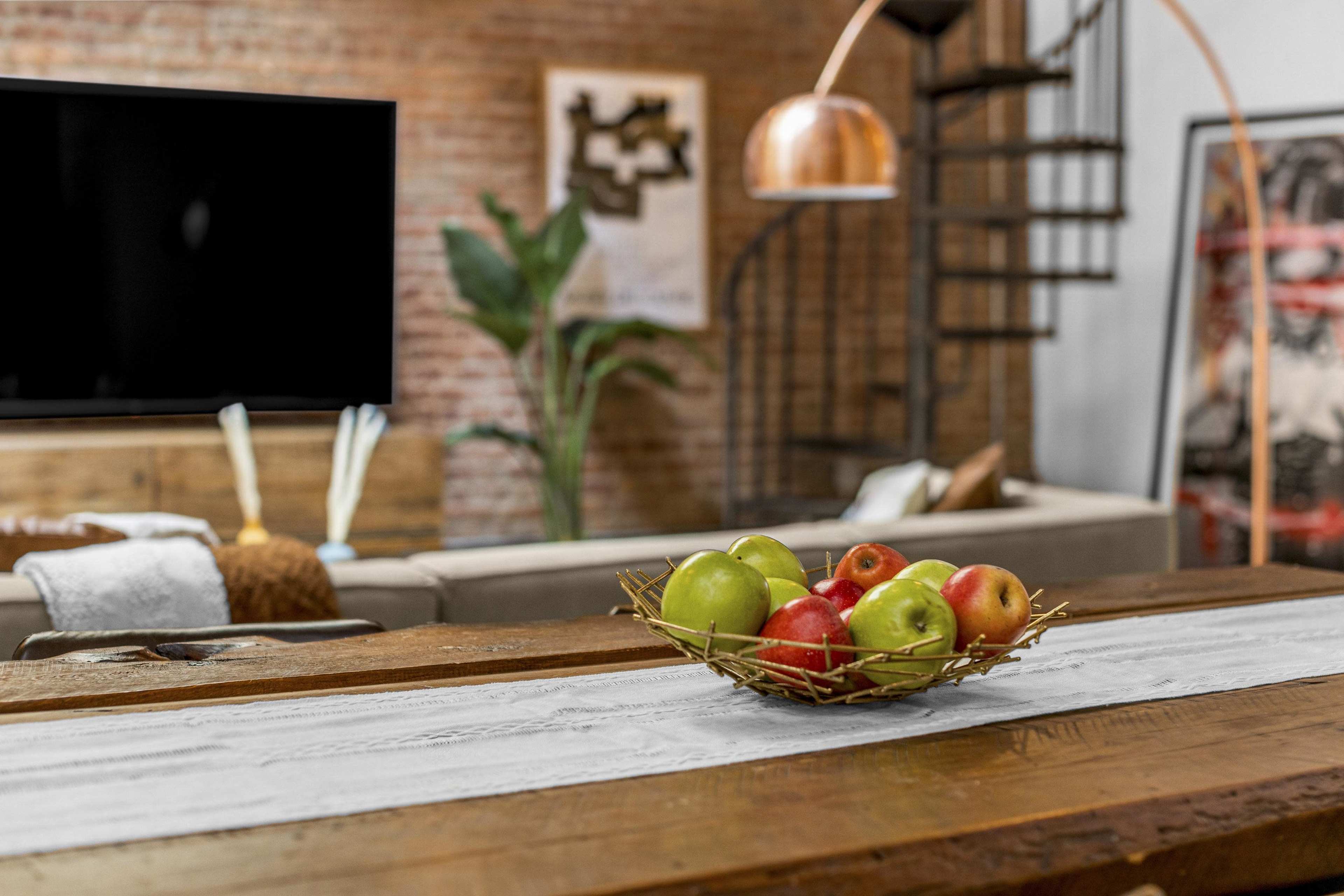 A bowl of apples sits on a wooden countertop in a modern living room with a spiral staircase and a large TV in the background.