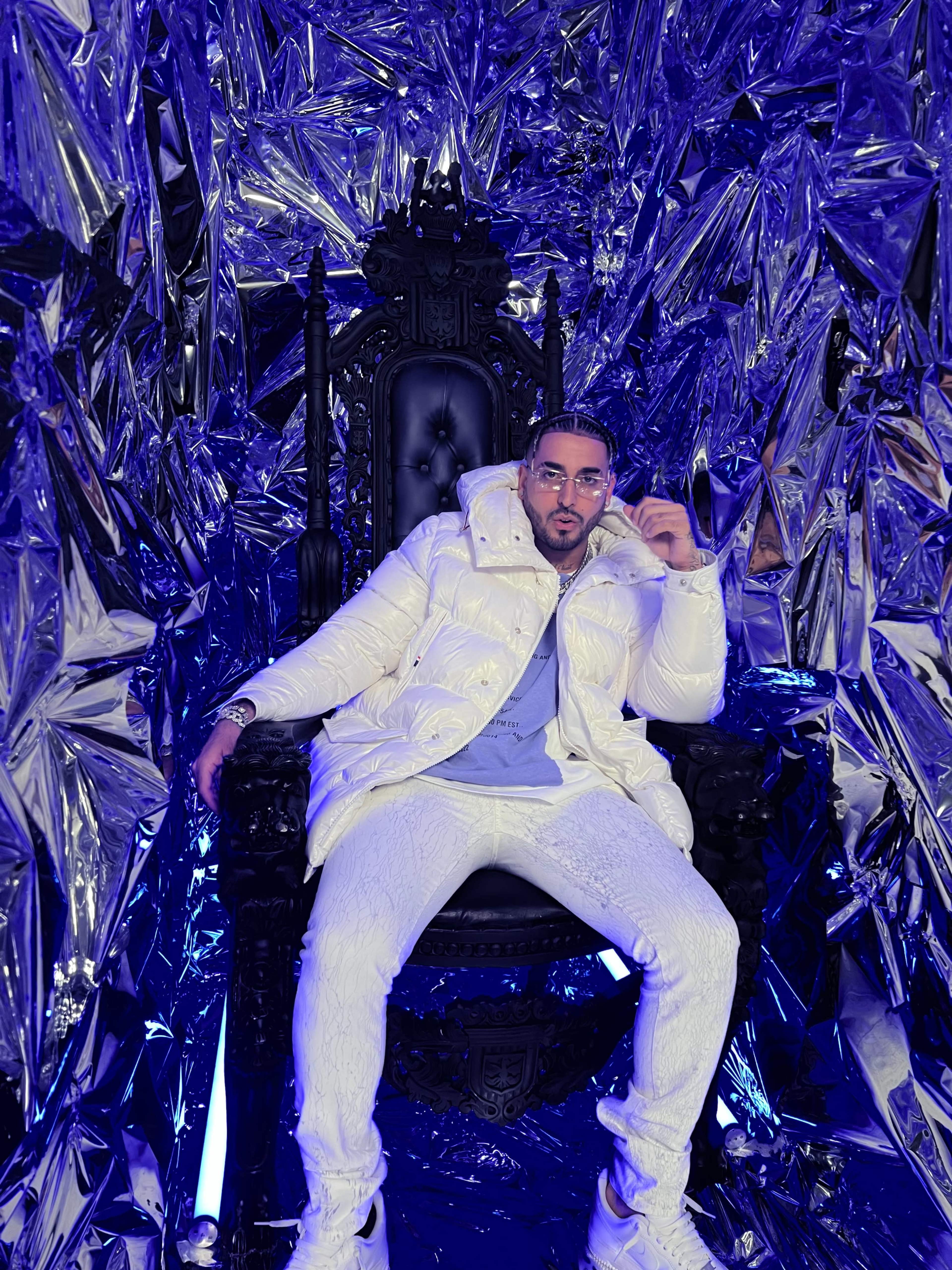 A person dressed in a white puffer jacket and matching pants sits on an ornate black throne surrounded by reflective, metallic decor.
