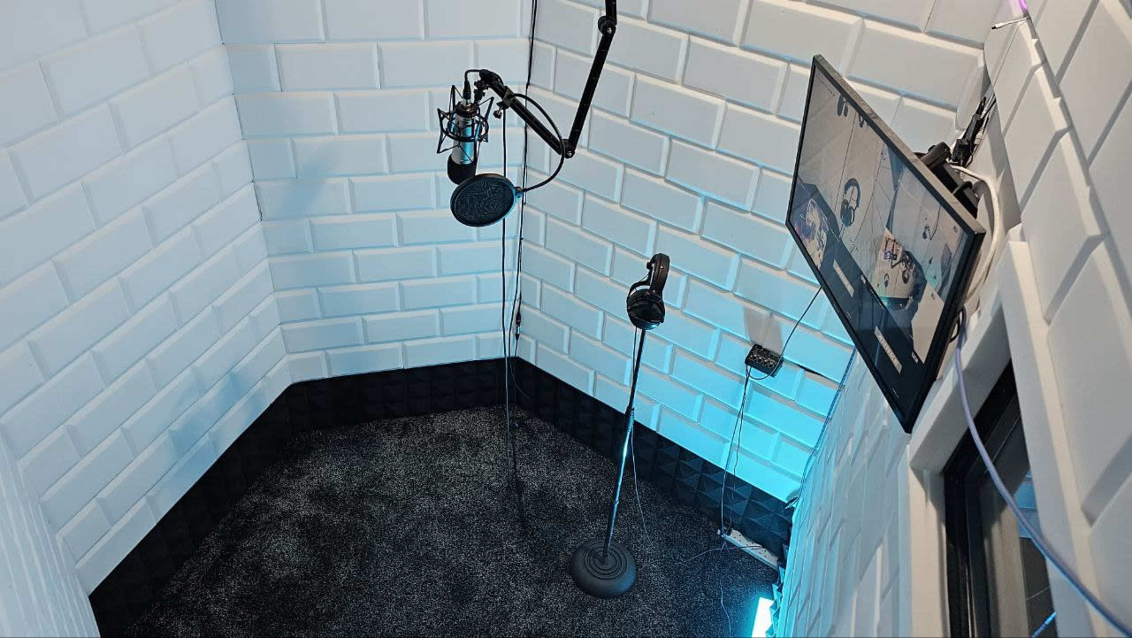 A small soundproof recording booth equipped with microphones, a monitor displaying video content, and walls covered in white sound-absorbing panels.