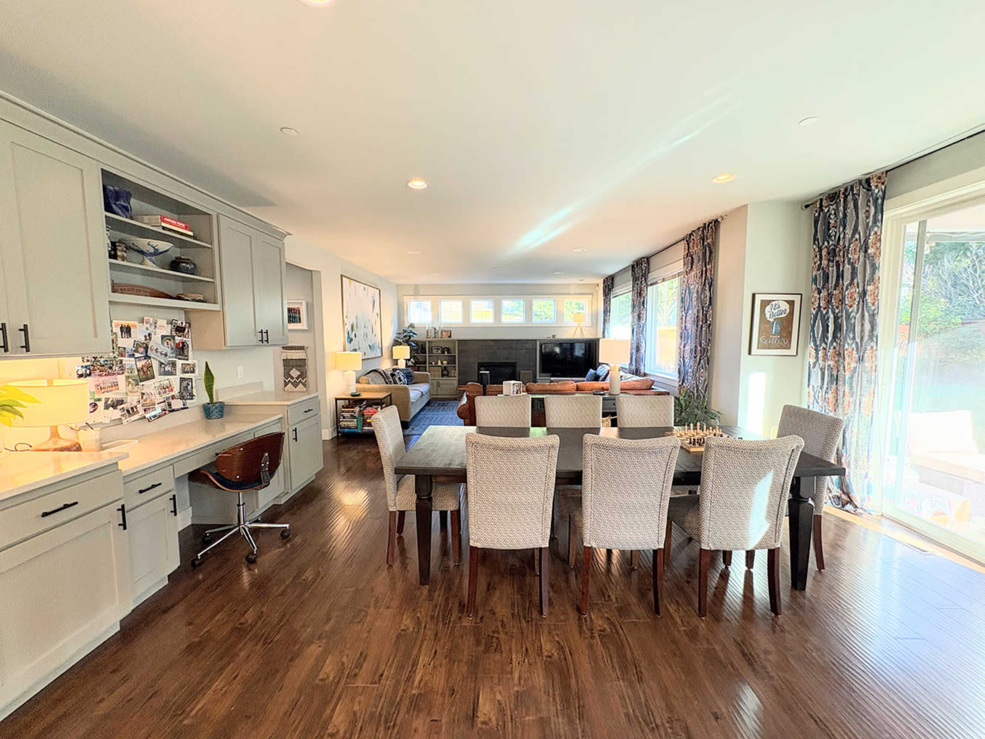 The image shows a modern, open-concept living and dining area featuring a long dining table surrounded by chairs, a kitchen area with cabinets, and large windows allowing natural light into the space.