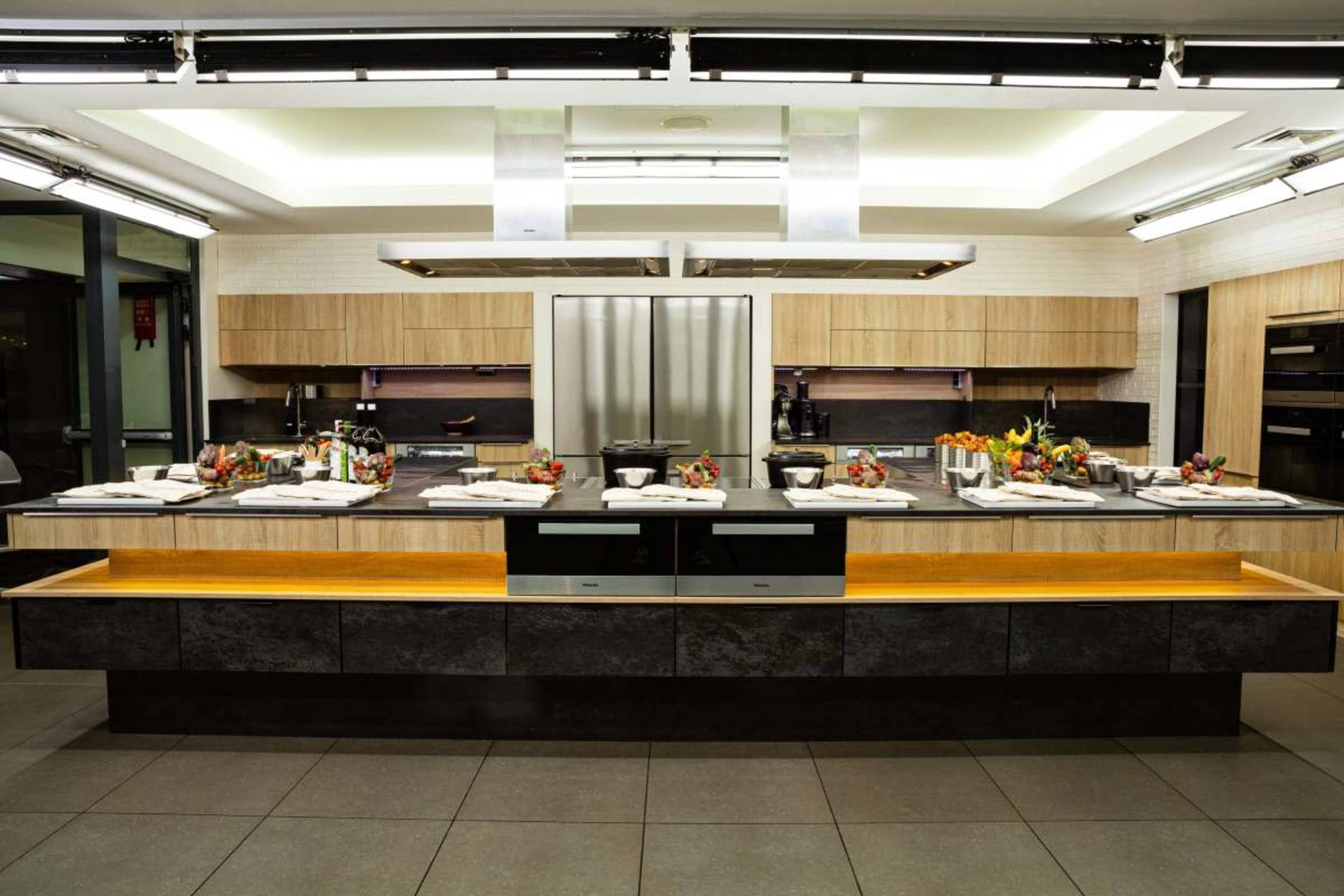 The image shows a modern kitchen with a central island, equipped with stainless steel appliances, wooden cabinetry, and a countertop decorated with bowls of fruits and cooking utensils.