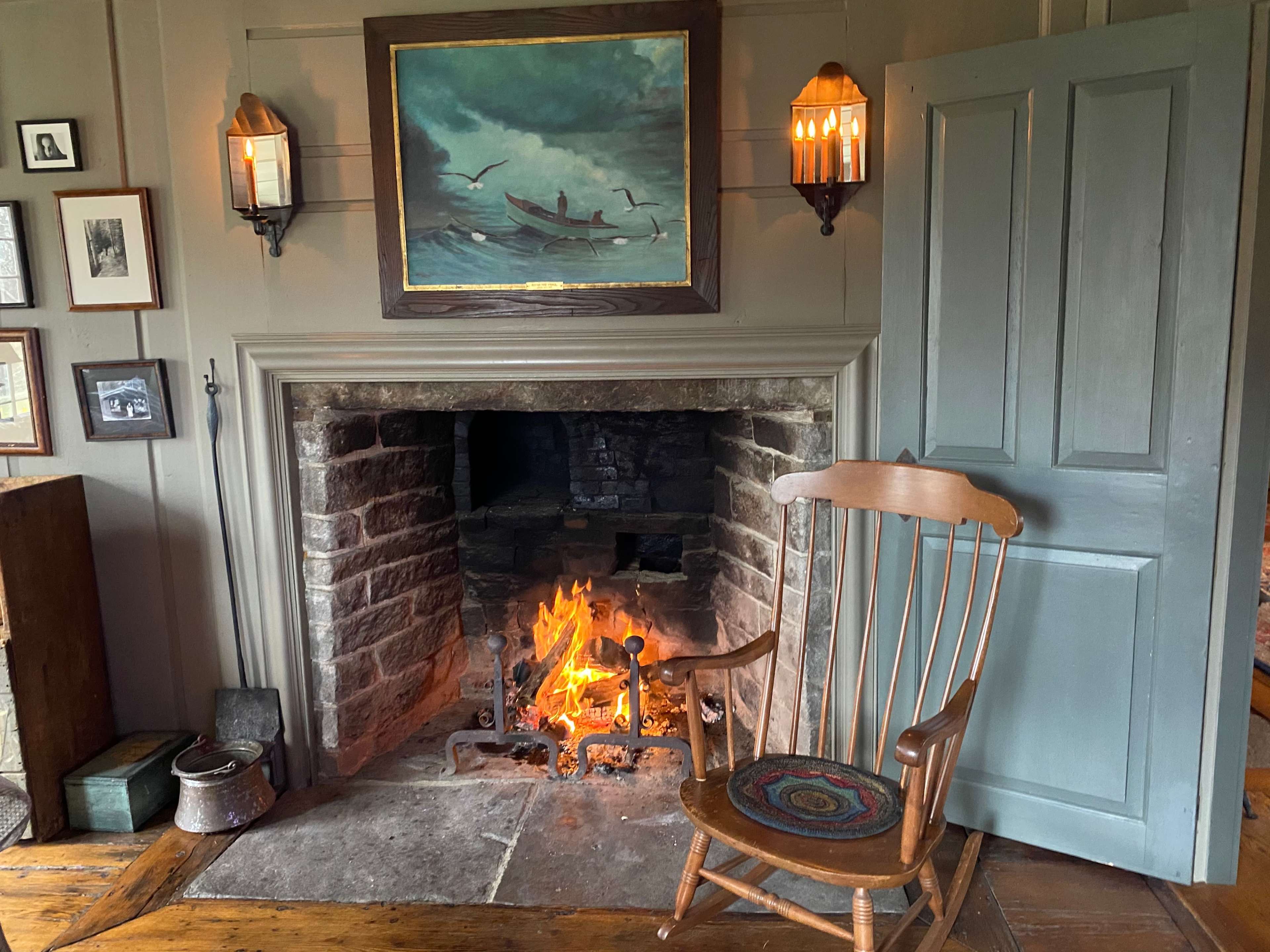 A cozy room with a stone fireplace, a rocking chair, a painting of boats, and wall-mounted lanterns.