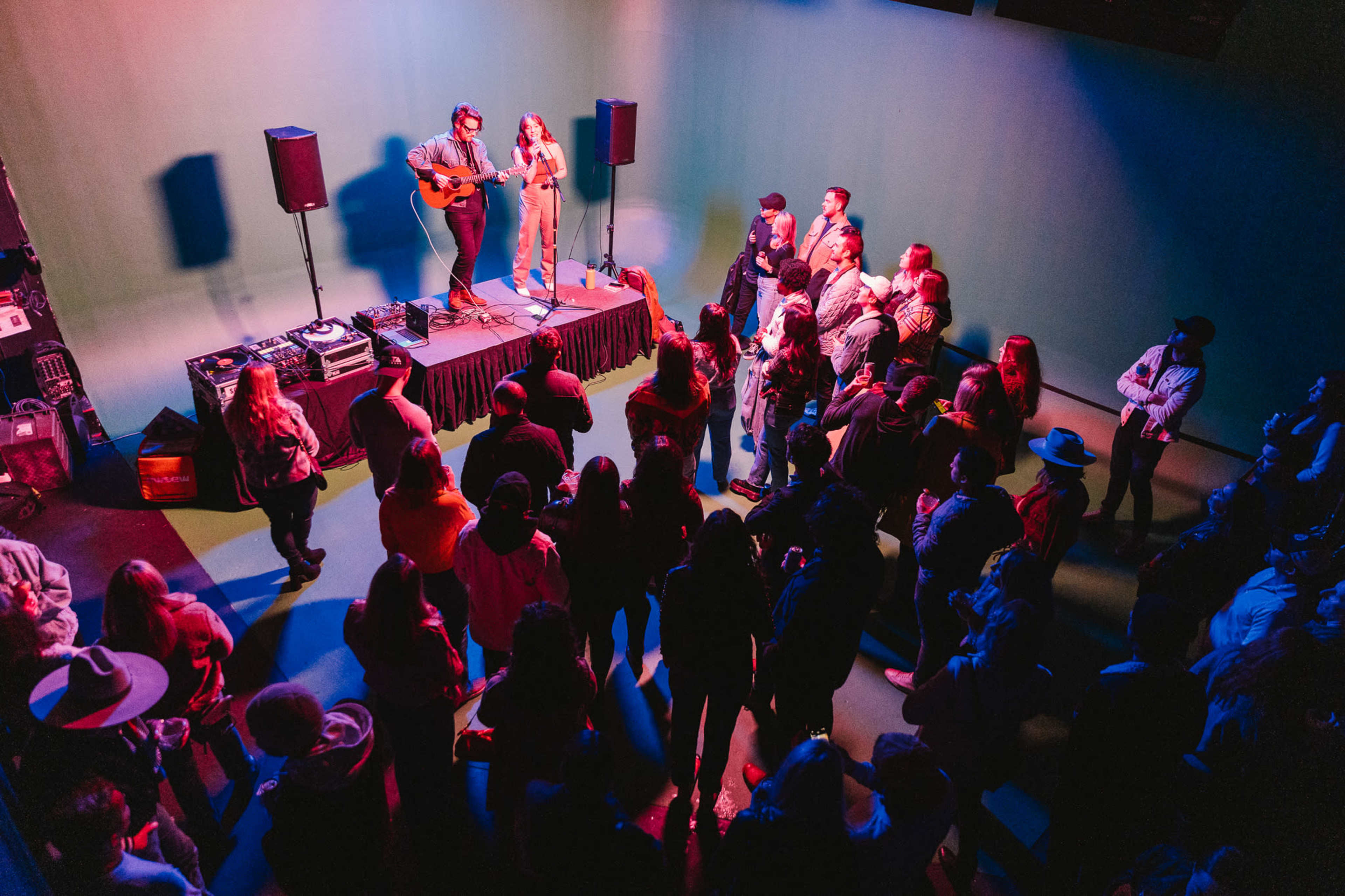 A crowd gathers around two musicians performing on a small stage with colorful lighting.